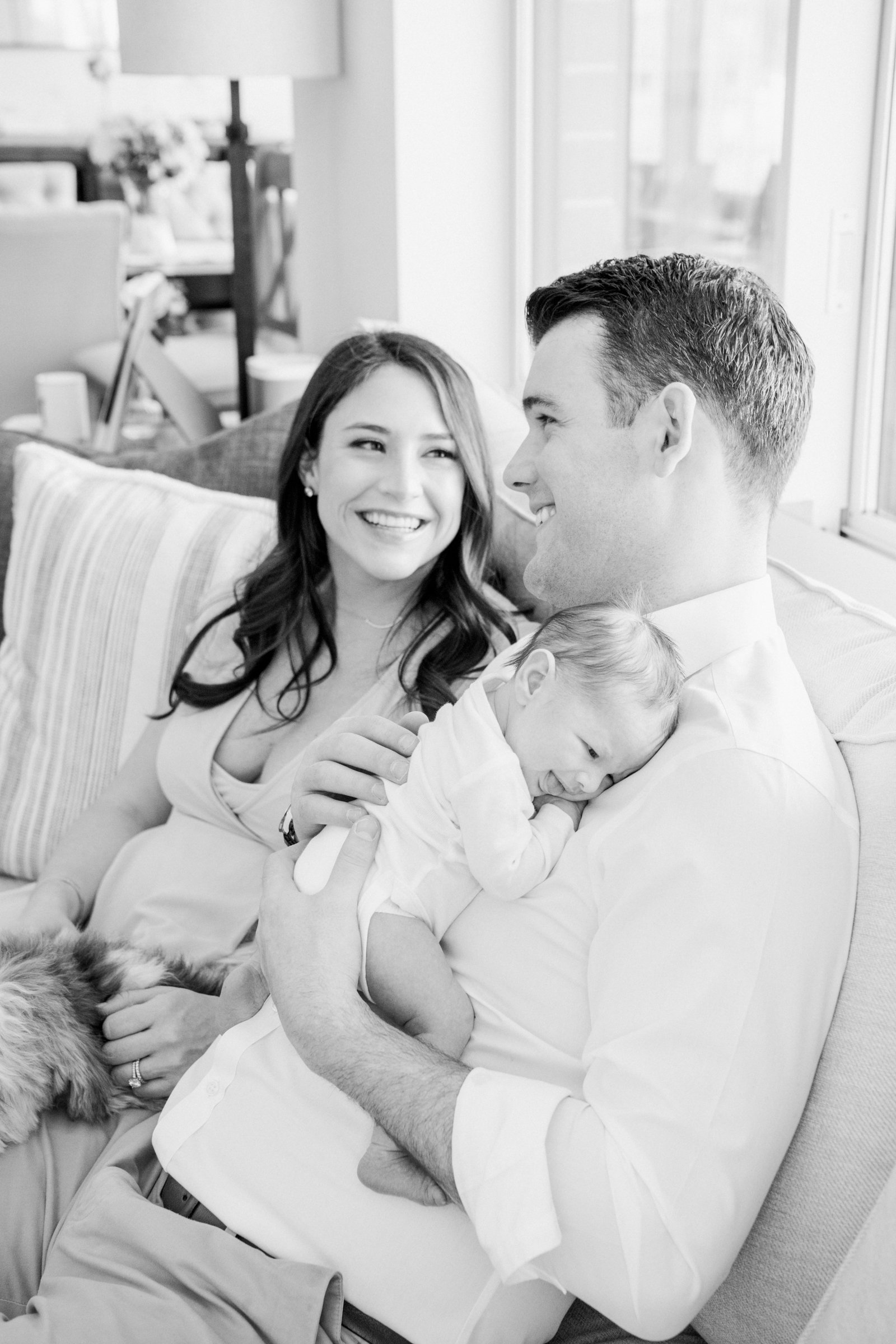  Father and mother sit on their couch during an in-home newborn session and smile at one another by Nicole Hawkins Photography in New Jersey. mother and father new parents fresh baby girl mother's love mother smile #nicolehawkinsphotography #nicoleha