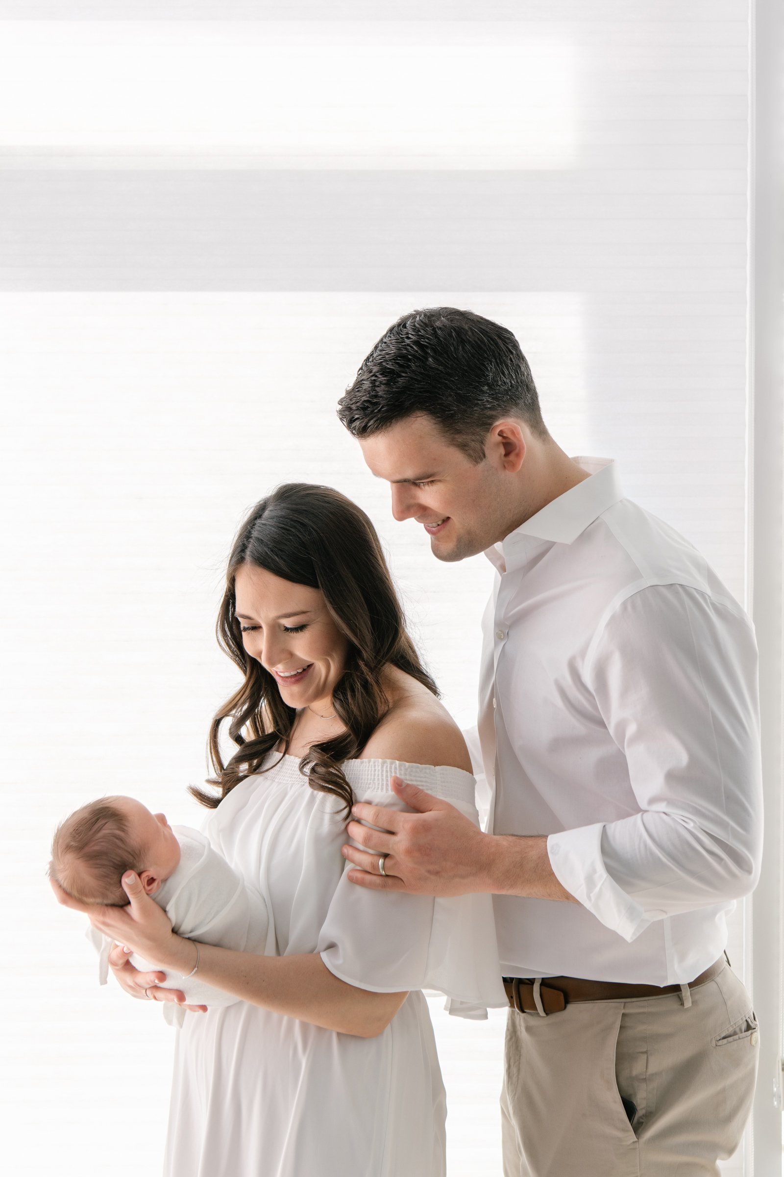  A mother and father stand in front of a bright light window and look down at their newborn baby girl in New Jersey by Nicole Hawkins Photography. family silhouette portraits newborn family portrait family of three pose ideas #nicolehawkinsphotograph