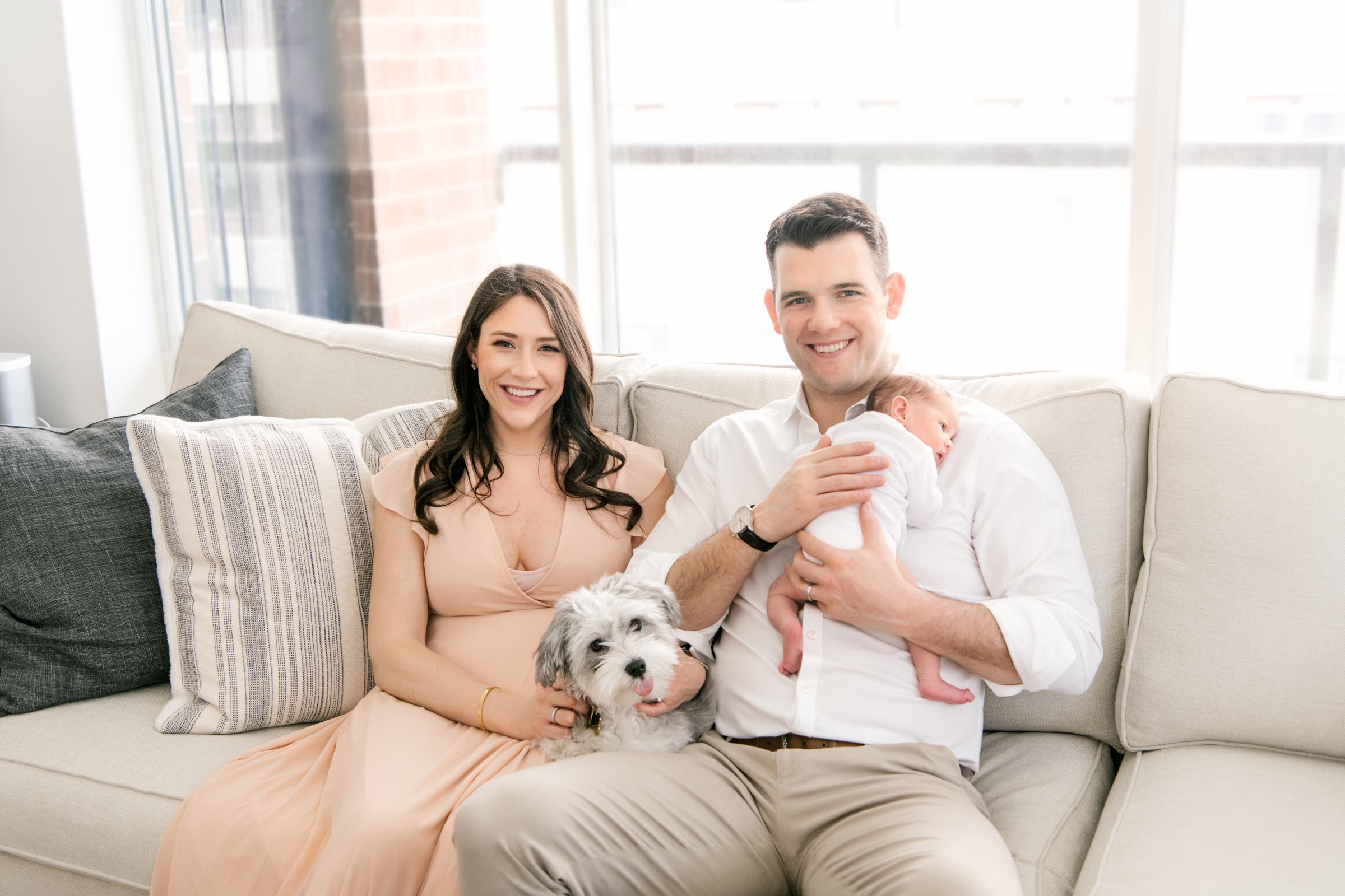  Family of four sitting on a couch with a baby on her father's shoulder with a puppy on the mother's lap by Nicole Hawkins Photography in Hoboken. family with puppy and baby in-home session family inspiration pose for in-home #nicolehawkinsphotograph