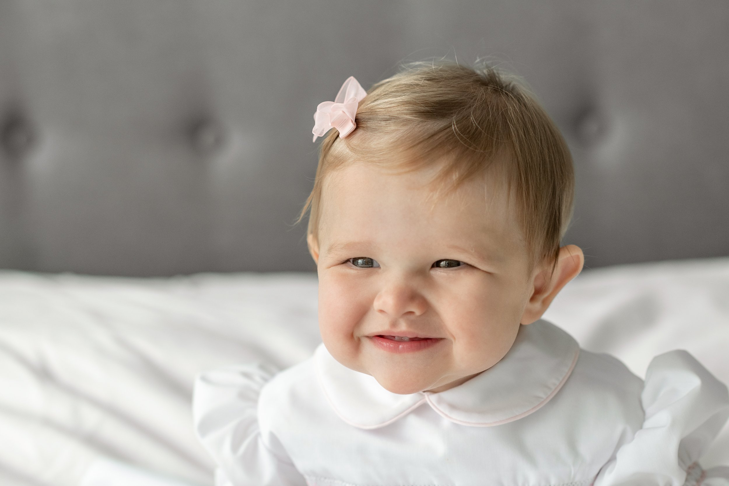  Nicole Hawkins Photography captures a portrait of a little girl smiling with a peter pan collar. NJ photographers little girl with bow white dress #nicolehawkinsphotography #newjerseyphotographers #familyphotographer #6monthportraits #NJfamilyphotos