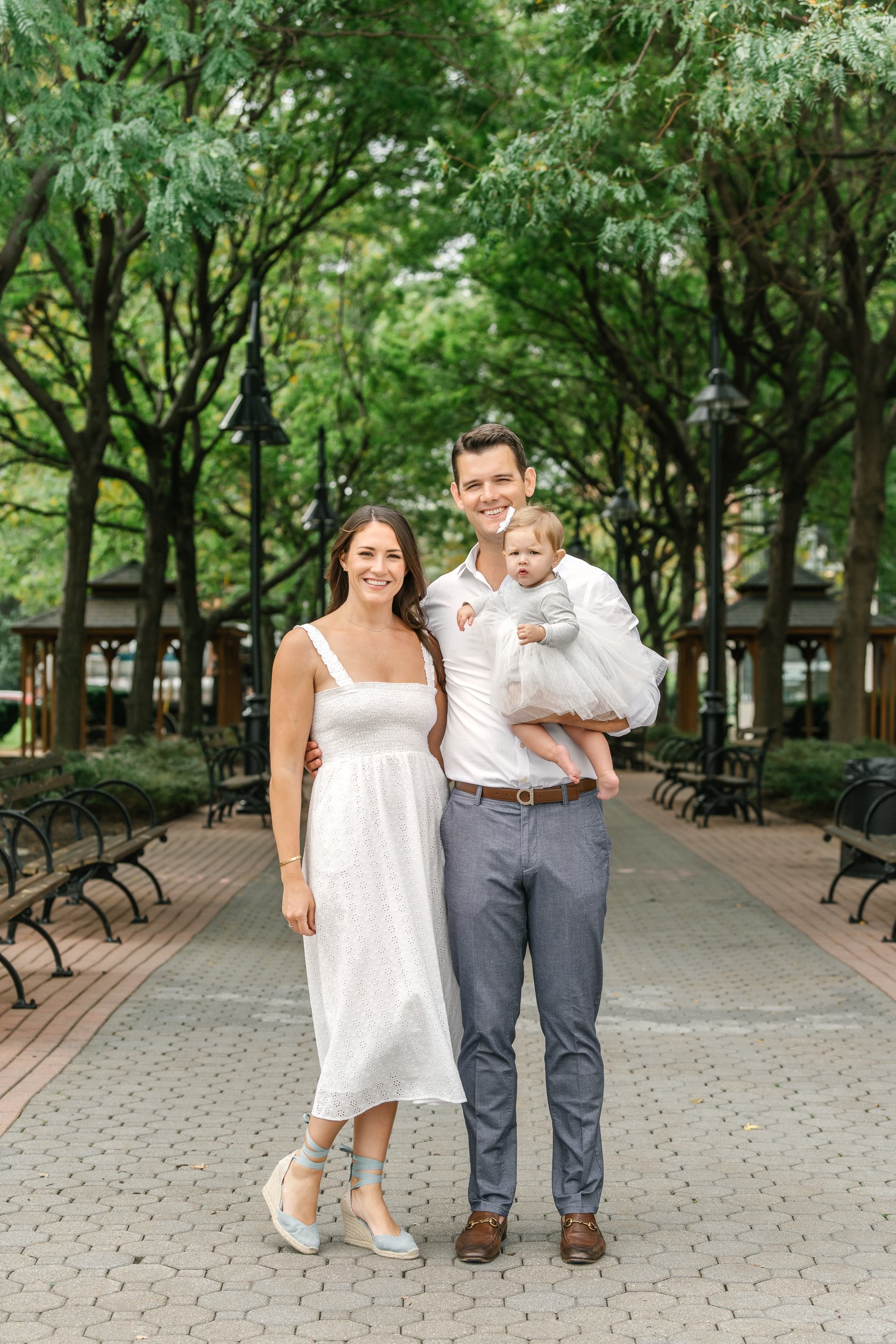  On a green paved park pathway, a young family takes portraits all in white with Nicole Hawkins Photography. park family pic family with a daughter #nicolehawkinsphotography #newjerseyphotographers #familyphotographer #6monthportraits #NJfamilyphotos