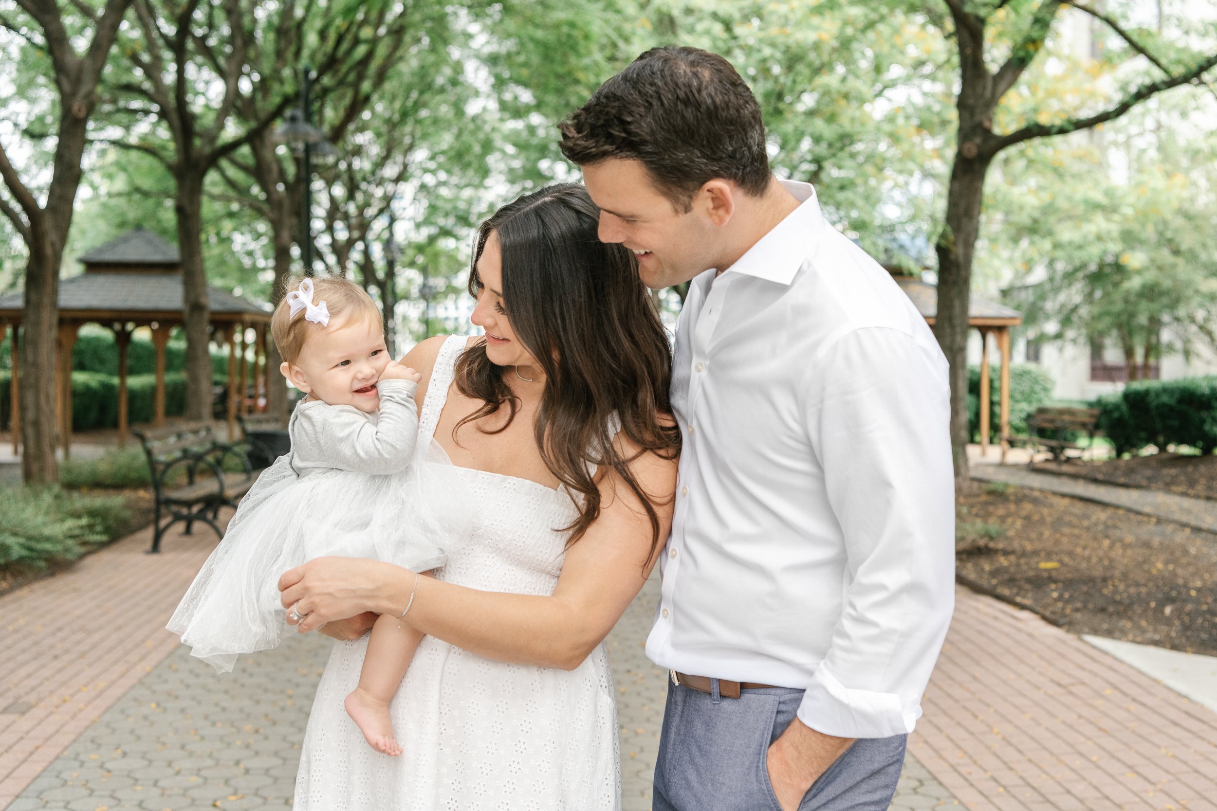  Mother and father look lovingly at their infant daughter in a white dress by Nicole Hawkins Photography. white dress family poses #nicolehawkinsphotography #newjerseyphotographers #familyphotographer #6monthportraits #NJfamilyphotos 