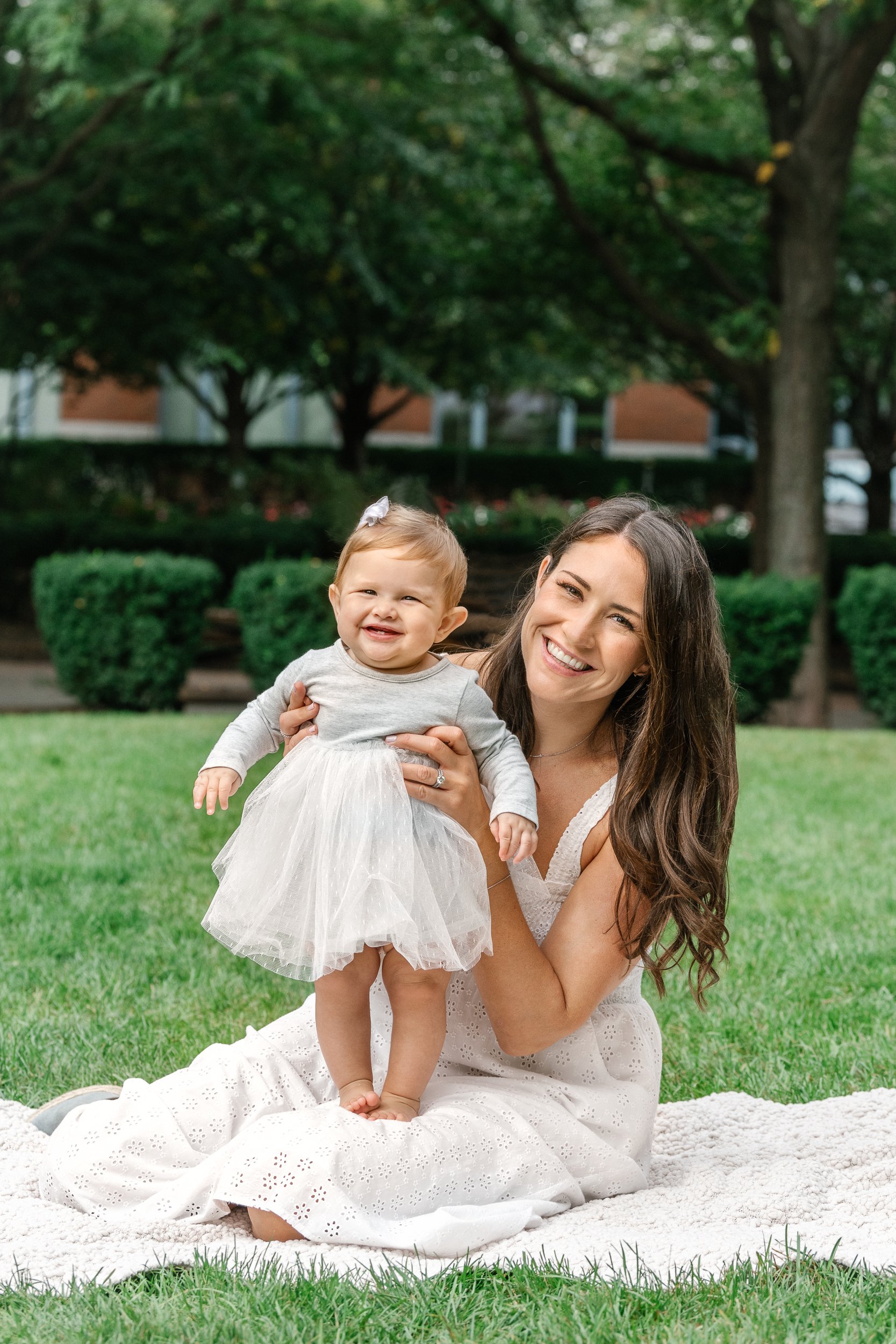  Nicole Hawkins captures a baby girl with a big smile standing on her mommy's lap in Hoboken. eyelet white dress mother and baby picnic blanket #nicolehawkinsphotography #newjerseyphotographers #familyphotographer #6monthportraits #NJfamilyphotos 