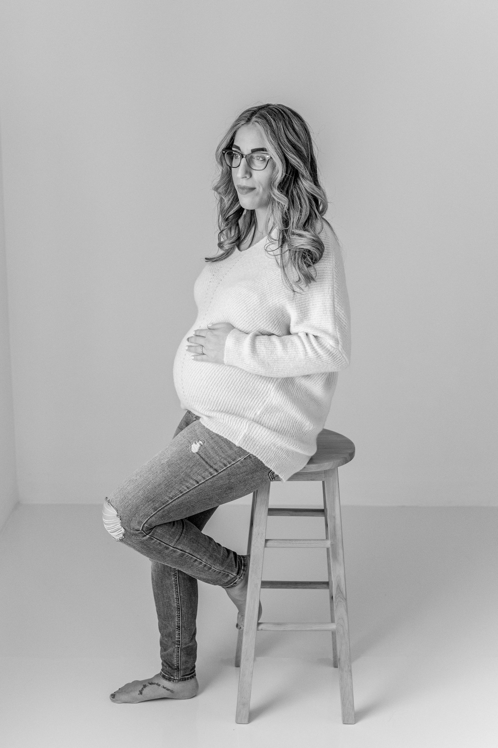  A pregnant woman sits on a stool during a studio session with modern chic style by Nicole Hawkins Photography. modern chic maternity pics NJ #NicoleHawkinsPhotography #maternitysession #NJmaternityphotographer #NewJerseyphotographers #maternityphoto