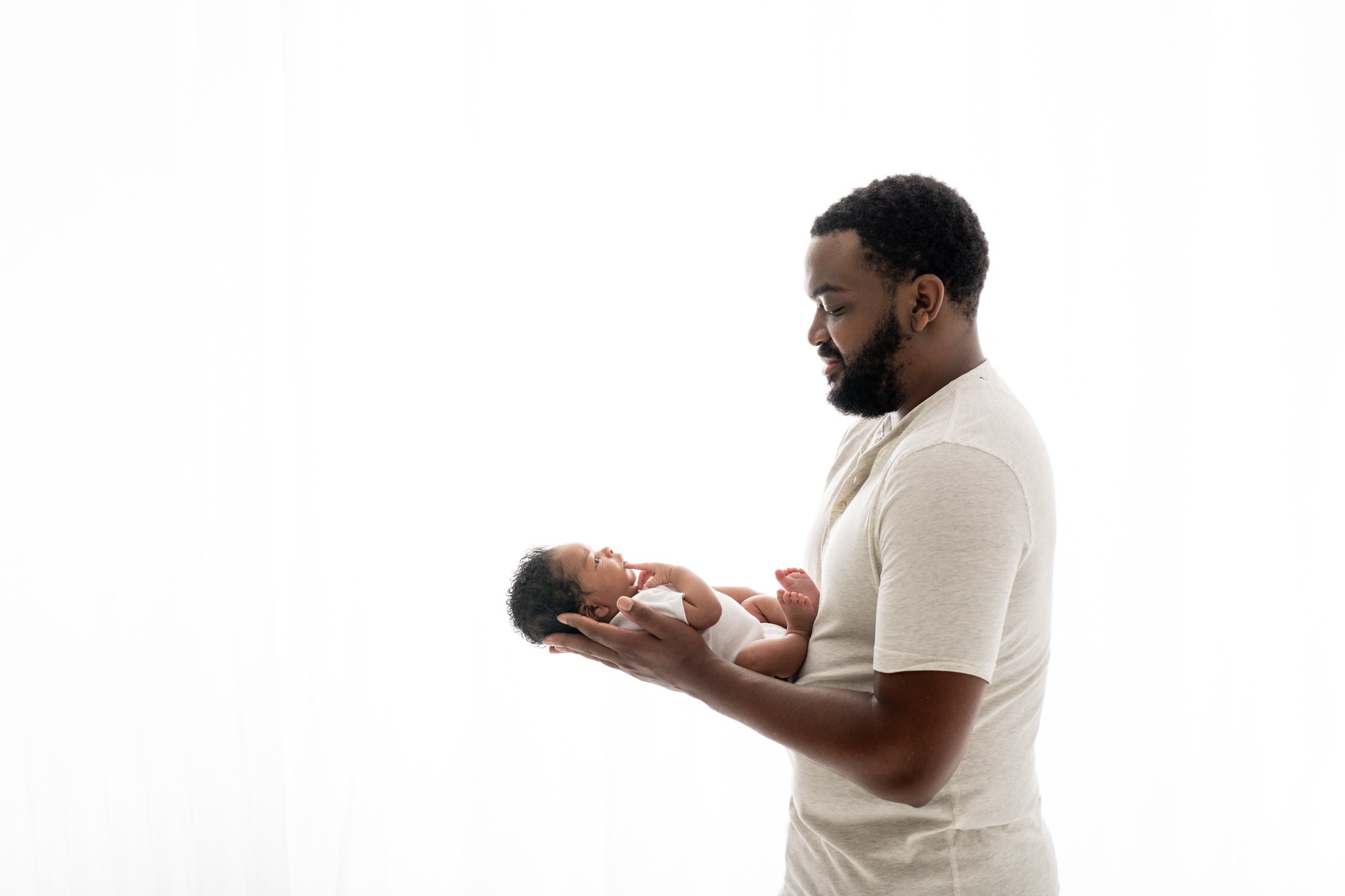  In front of a bright white window, a father holds his son in his arms by Nicole Hawkins Photography in New Jersey. father at newborn jr and dad #nicolehawkinsphotography #NJbabyphotography #newbornportraits #NewJerseyStudioPhotography #newbornsessio