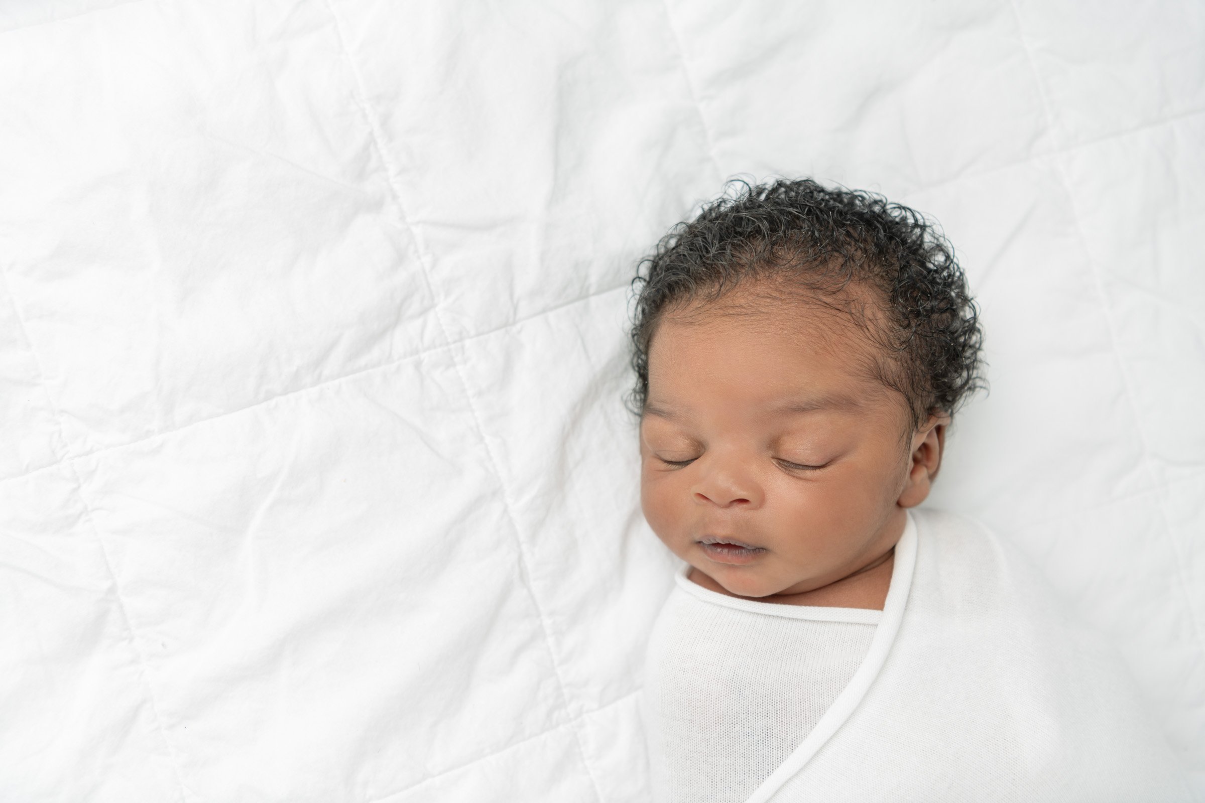  New Jersey photographer Nicole Hawkins Photography captures a swaddled sleeping baby on a white blanket. swaddled newborn curly black hair #nicolehawkinsphotography #NJbabyphotography #newbornportraits #NewJerseyStudioPhotography #newbornsession 