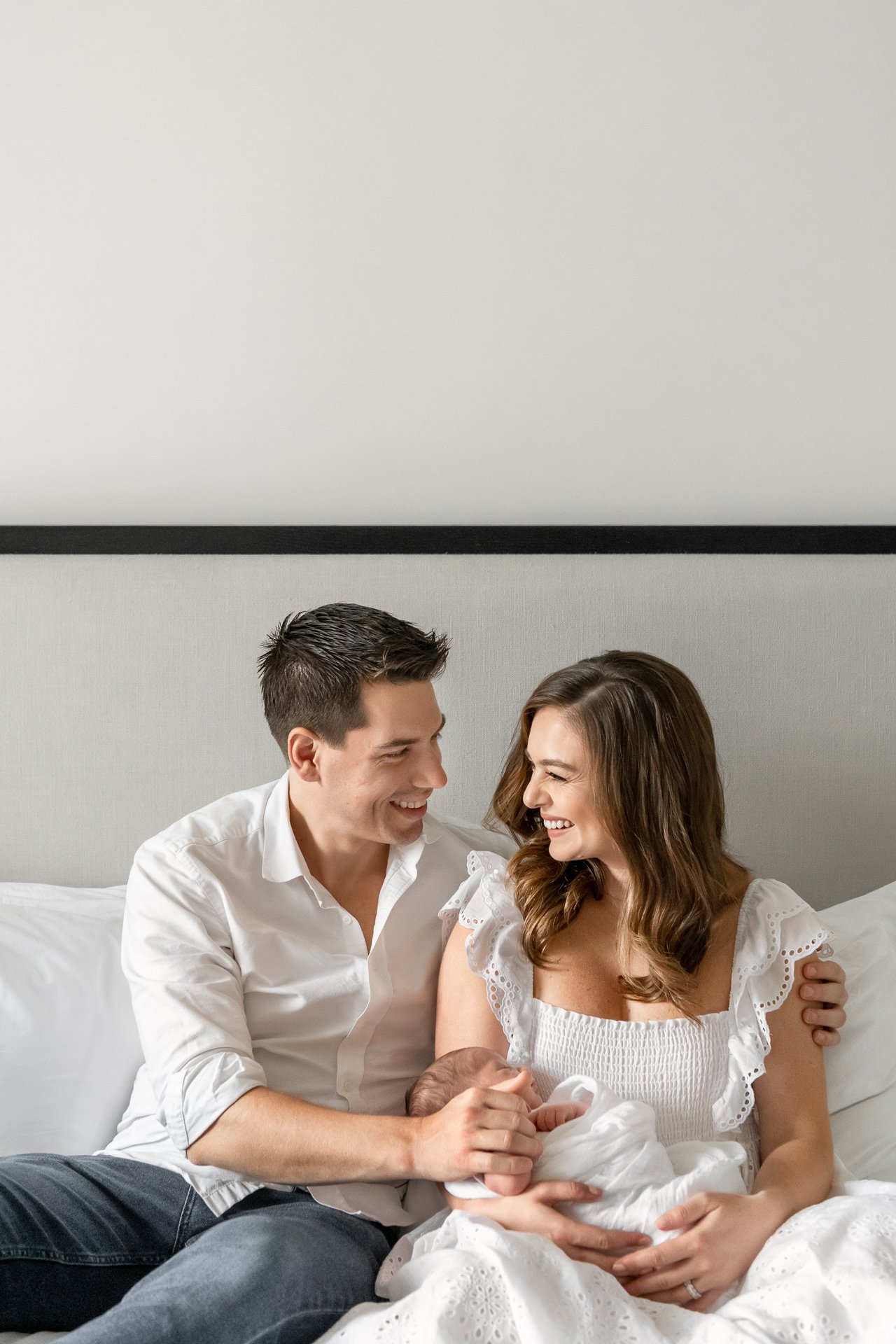  On a bed, a husband and wife smile at each other while holding their baby by Nicole Hawkins Photography. family portraits sitting on a bed #nicolehawkinsphotography #NYCbabyphotography #newbornportraits #NewYorkStudioPhotography #newbornsession 