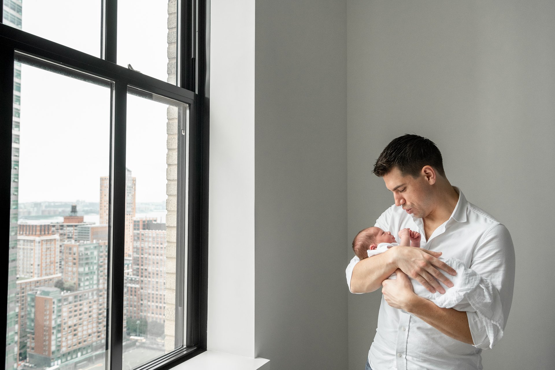  With the New York skyline out the window, a father rocks his baby by Nicole Hawkins Photography. modern newborn family portraits NYC #nicolehawkinsphotography #NYCbabyphotography #newbornportraits #NewYorkStudioPhotography #newbornsession 