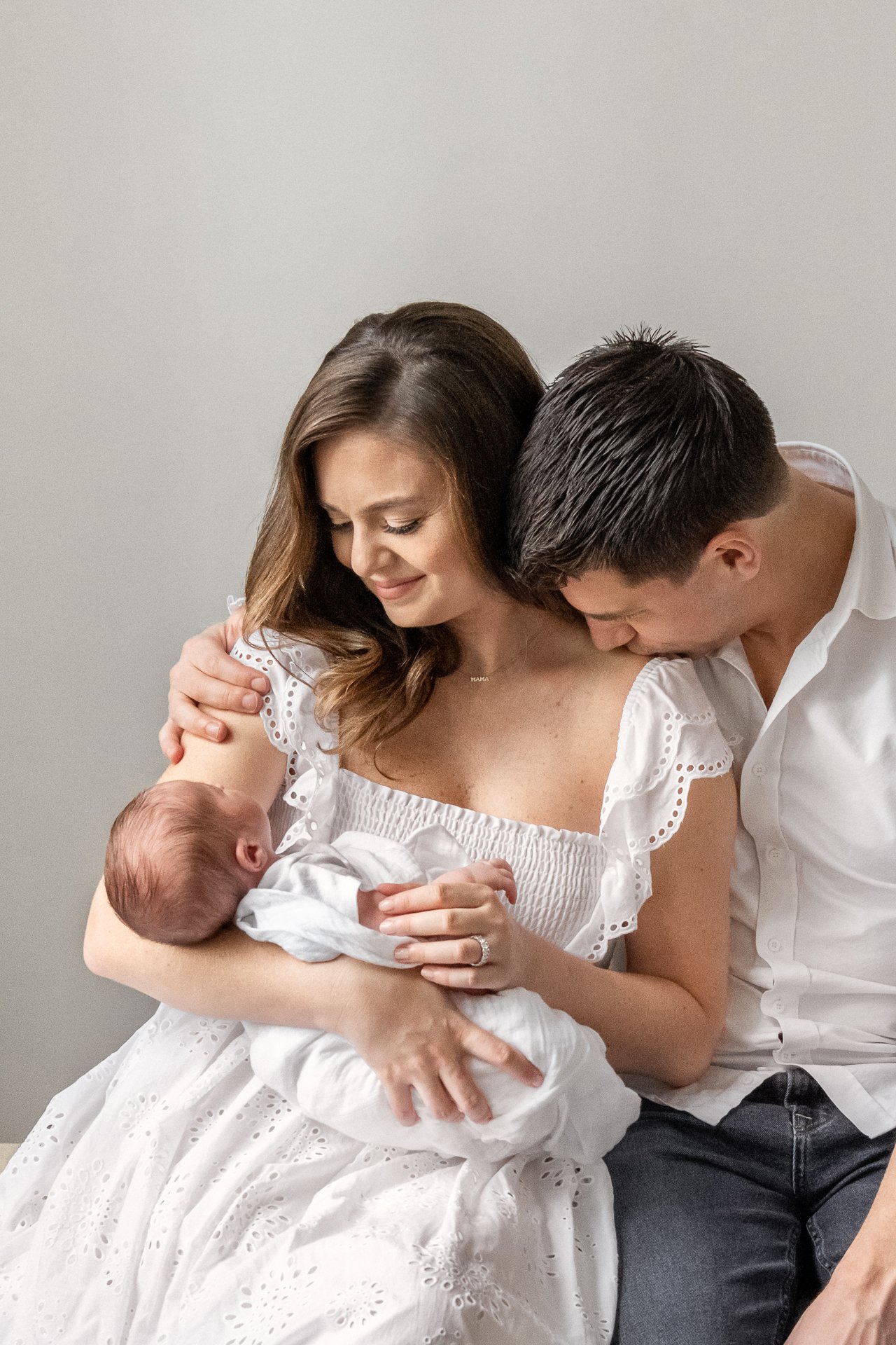  NY photographer Nicole Hawkins Photography captures a father kissing his wife's shoulder as she snuggles the baby. new parents family of three #nicolehawkinsphotography #NYCbabyphotography #newbornportraits #NewYorkStudioPhotography #newbornsession 