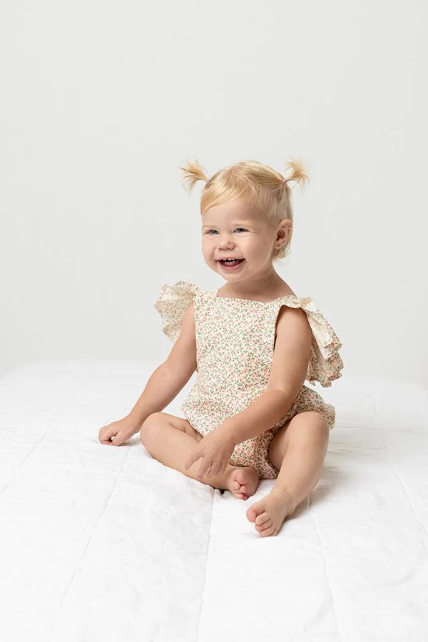  A toddler girl sitting on a bed with a big smile by Nicole Hawkins Photography. studio baby session baby girl studio session ideas #nicolehawkinsphotography #NJstudioportraits #babyportraits #babygirlportraits #NJbabyphotographers #NJphotographers 