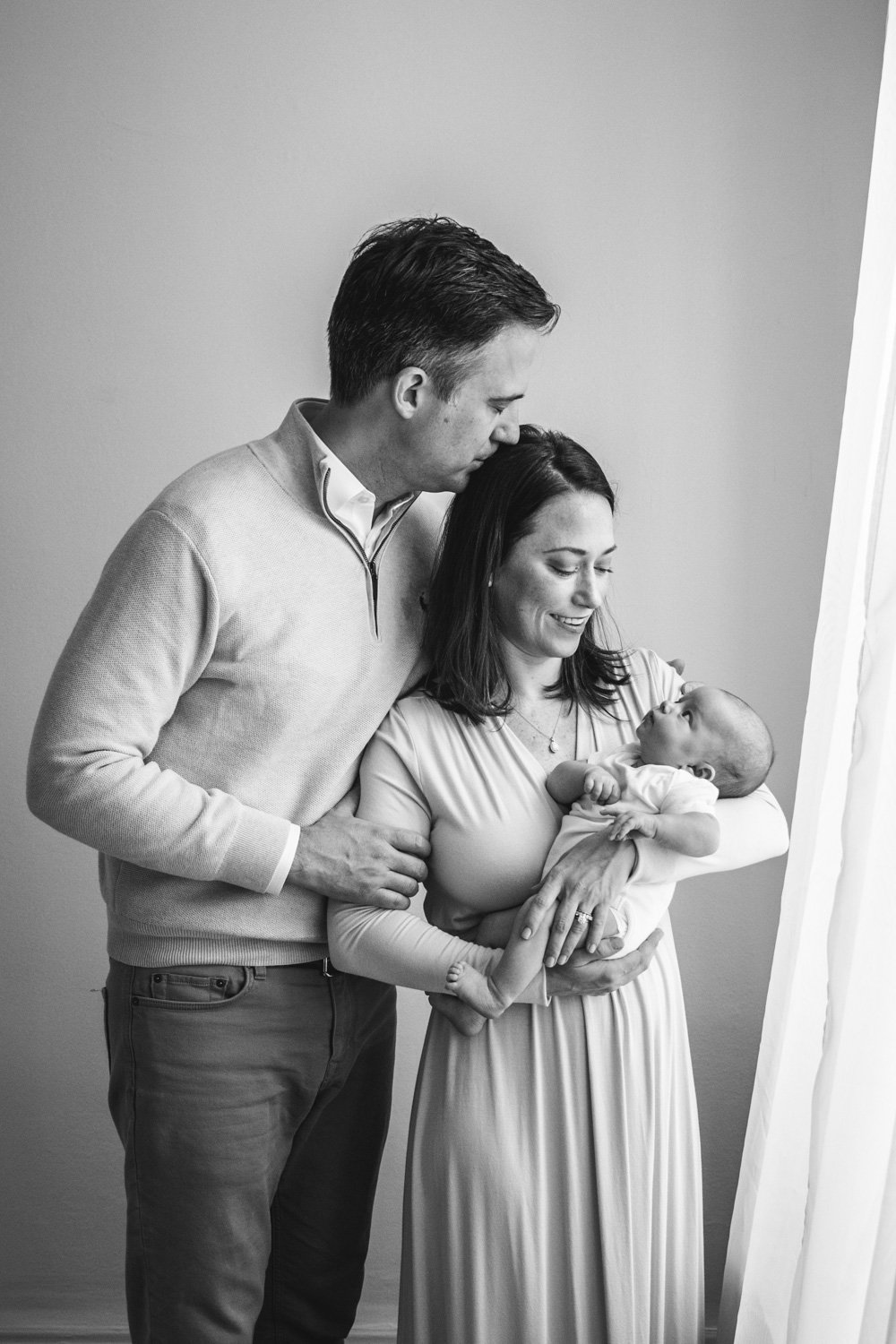 A black and white portrait of a father kissing his wife while she holds the newborn by Nicole Hawkins Photography. newborn studio session #nicolehawkinsphotography #NJstudionewborns #newborn #studionewborns #NJnewbornphotographers #NJphotographers 