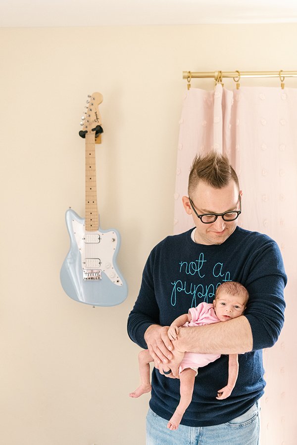  A father holds his newborn next to a baby blue electric guitar on the wall by Nicole Hawkins Photography. guitar in nursery mod nursery #nicolehawkinsphotography #NJfamilyphotographer #inhomenewbornsession #nicolehawkinsnewborns #NJnewbornphotograph