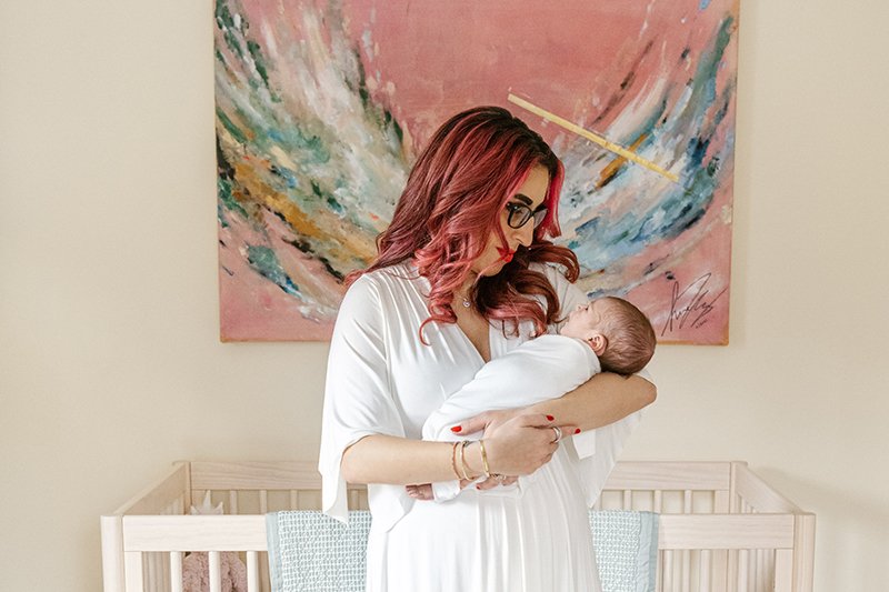 With an abstract painting behind her, a mother smiles at her baby by Nicole Hawkins Photography. abstract nursery wall art newborns NJ #nicolehawkinsphotography #NJfamilyphotographer #inhomenewbornsession #nicolehawkinsnewborns #NJnewbornphotography