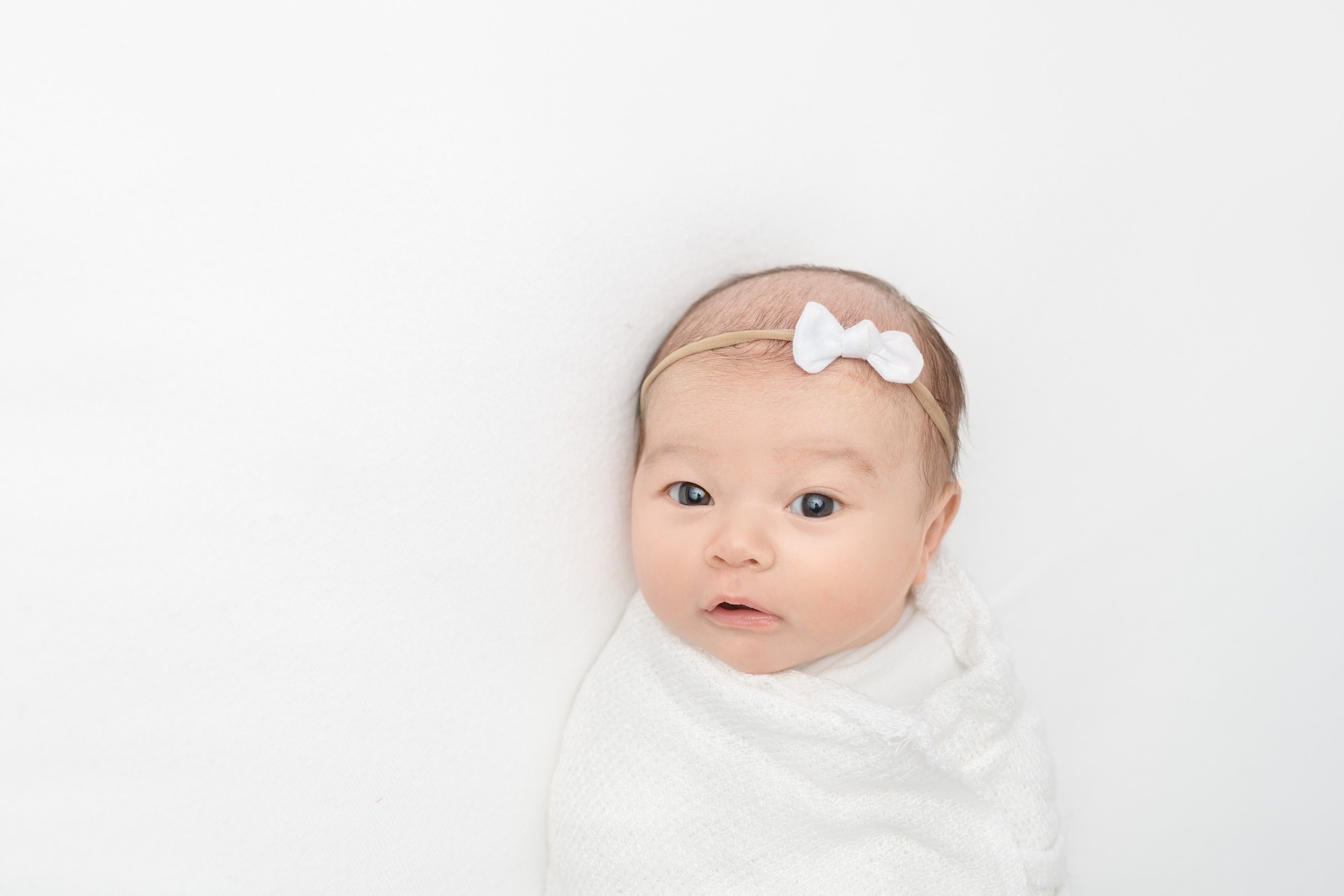  A baby swaddled in white wearing a bow was captured in a studio in Maplewood by Nicole Hawkins Photography. baby girl swaddle in white #NicoleHawkinsPhotography #studionewbornportraits #NJstudiophotographer #NJnewborns #NicoleHawkinsNewborns 
