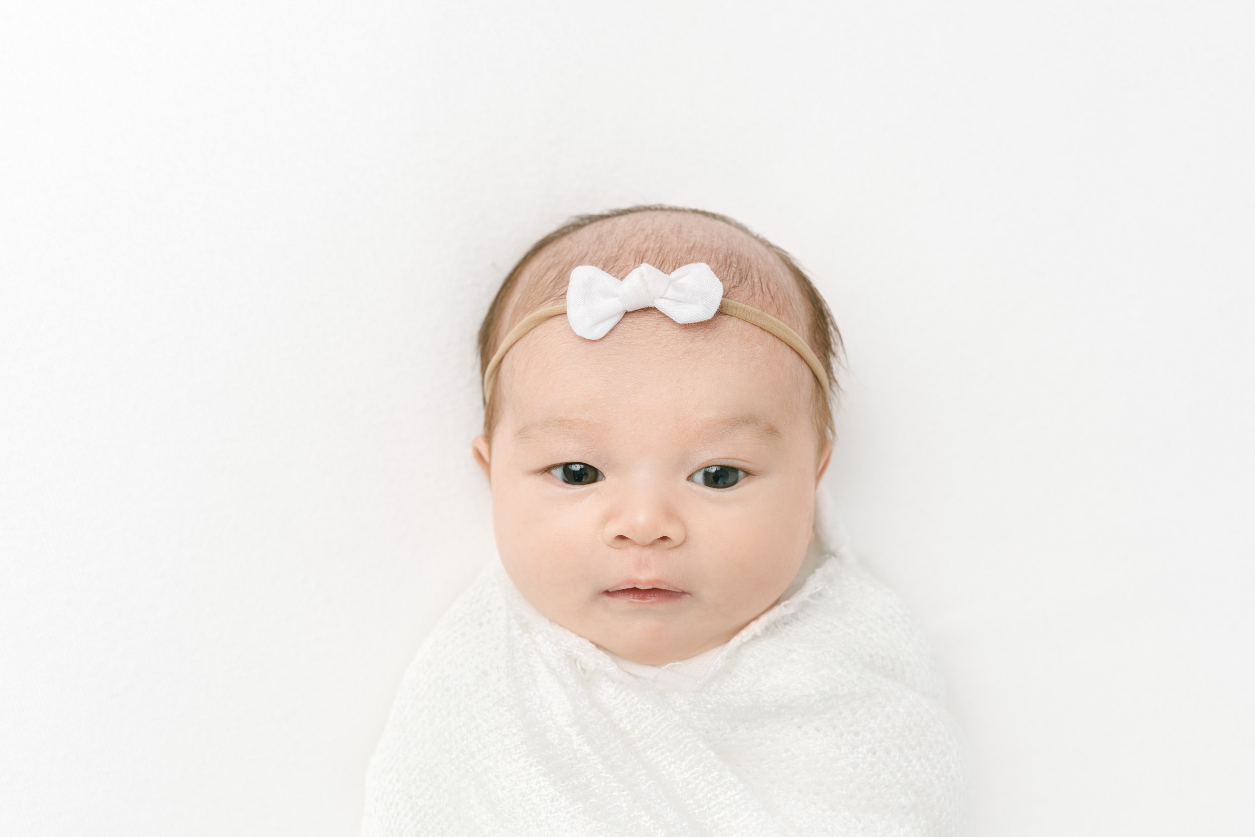  Baby girl with eyes wide open wearing a white bow captured by NJ newborn photographer Nicole Hawkins Photography. baby with eyes open white bow #NicoleHawkinsPhotography #studionewbornportraits #NJstudiophotographer #NJnewborns #NicoleHawkinsNewborn
