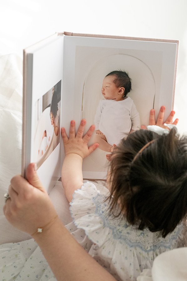  A one-year-old little girl looks at a picture book with a newborn portrait of herself by Nicole Hawkins Photography.  a baby then now baby girl #nicolehawkinsphotography #nicolehawkinsbirthday #nicolehawkinsportraits #NJstudiophotography #1stbirthda