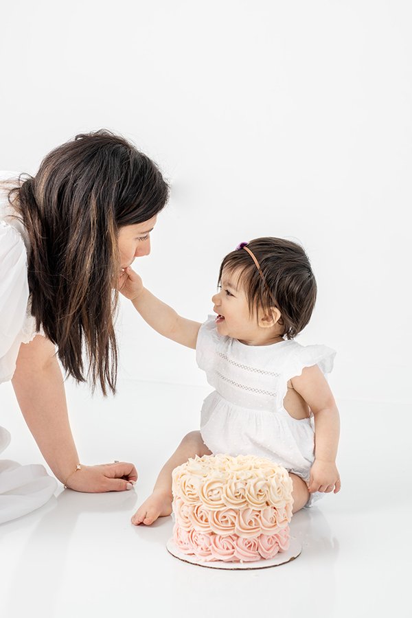  Mother talks to her baby while she eats her smash cake in a studio in NJ by Nicole Hawkins Photography. pink cake first birthday #nicolehawkinsphotography #nicolehawkinsbirthday #nicolehawkinsportraits #NJstudiophotography #1stbirthday 