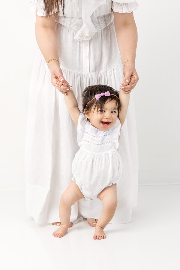  A mother holds her little girl's hands as the girl bounces around by Nicole Hawkins Photography. toddler poses toddler New Jersey toddlers #nicolehawkinsphotography #nicolehawkinsbirthday #nicolehawkinsportraits #NJstudiophotography #1stbirthday 