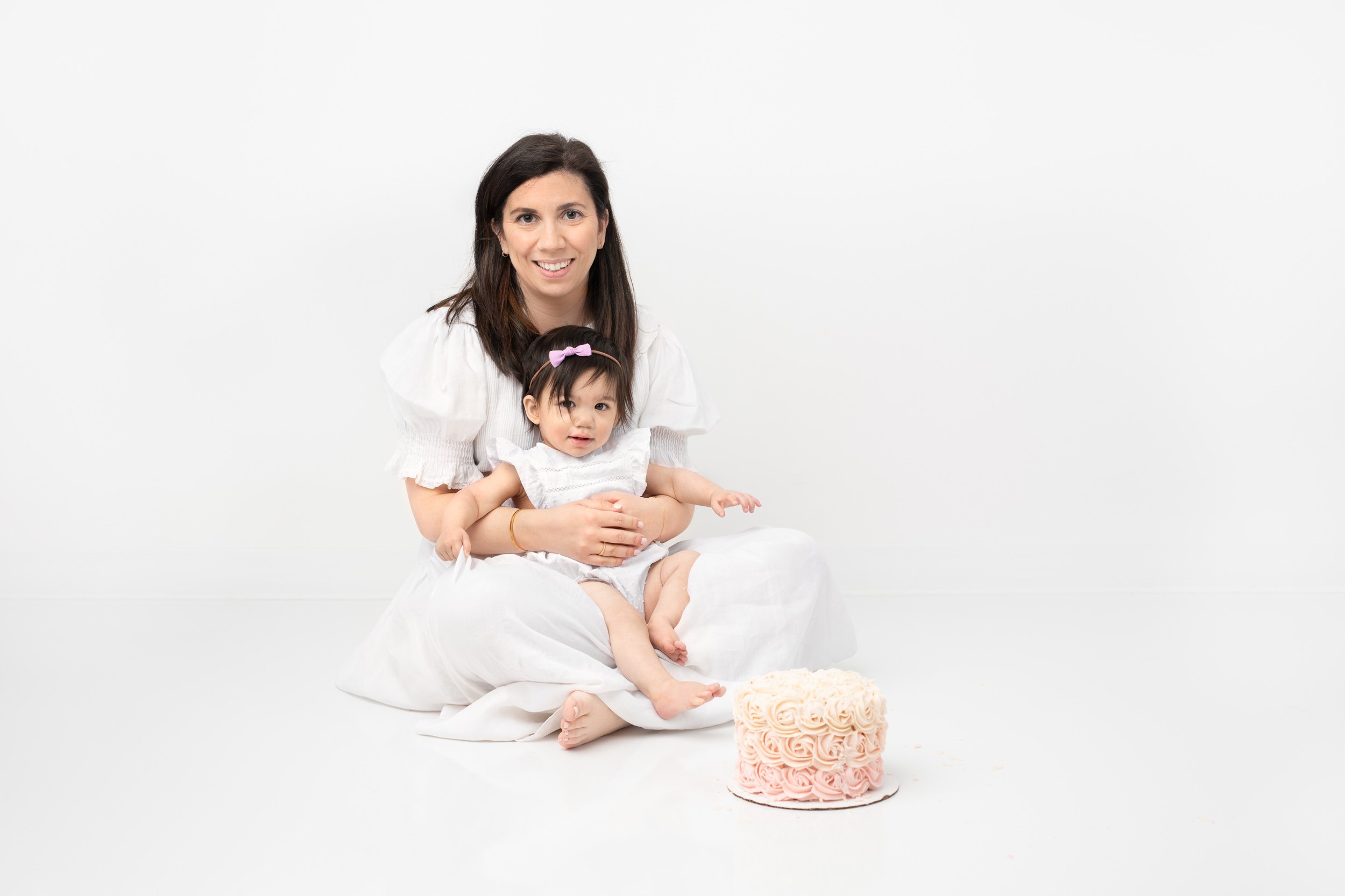 Baby girl sits in her mom's lap on a studio floor next to her birthday cake by Nicole Hawkins Photography. motherhood milestone pics #nicolehawkinsphotography #nicolehawkinsbirthday #nicolehawkinsportraits #NJstudiophotography #1stbirthday 