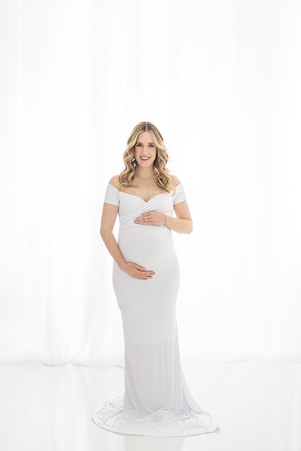  In a white floor-length gown, a pregnant woman smiles at herself by Nicole Hawkins Photography. white gown maternity family photographer NJ #nicolehawkinsphotography #nicolehawkinsmaternity #maternityportraits #NJstudiophotography #mommatobe 
