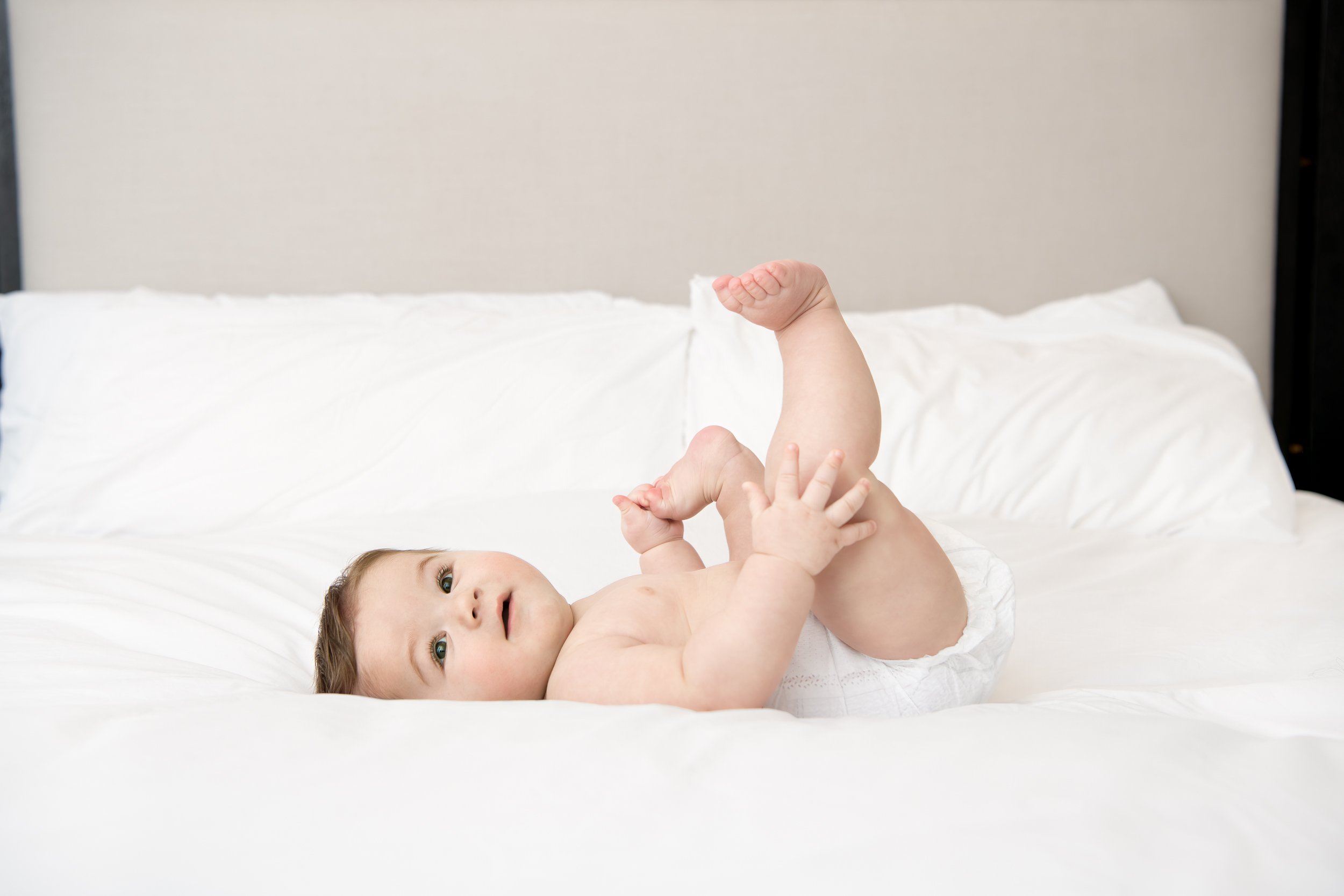  Baby laying on a white bed touching his toes by Nicole Hawkins Photography in New Jersey. baby toes baby portraits #NicoleHawkinsPhotography #NicoleHawkinsMilestones #BabyMilestoneSession #NJfamilyphotographer #Inhomephotography #babyportraits 