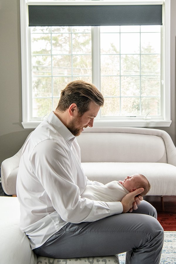  New Jersey newborn photographer Nicole Hawkins Photography captures a father holding his son in front of a bright window. baby boy #NewbornSession #NJfamilyphotographer #Inhomebabyphotography #newborn #NicoleHawkinsPhotography #NicoleHawkinsNewborns