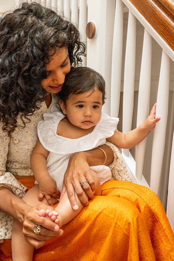  Mom with curly hair wearing a cream lace top and orange wide leg pants holds her baby on the stairs. #familyportraits #newjersyphotographers #familyoffour #njfamilyphotoographer #nicolehawkinsphotography #nicolehawkinsfamilies #glenridge #newjersey 