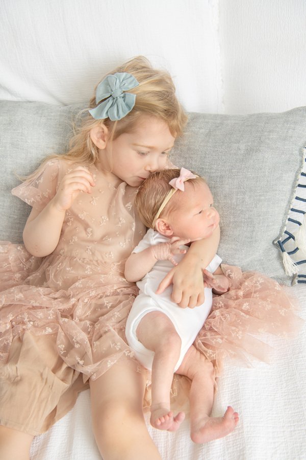 Toddler big sister wearing a pink tulle dress cuddles her newborn baby sister on a bed in their Chatham home. #nicolehawkinsphotography #secondchild #chatham #newjersey #inhomephotography #nicolehawkinsnewborns #newbornsession #njfamilyphotographer 