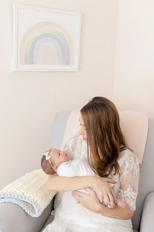  Second-time mom wearing a white lace dress holds her swaddled newborn daughter while rocking in the recliner. #inhomephotography #babynursery #nicolehawkinsphotography #chatham #newjersey #nicolehawkinsnewborns #newbornsession #njfamilyphotographer 