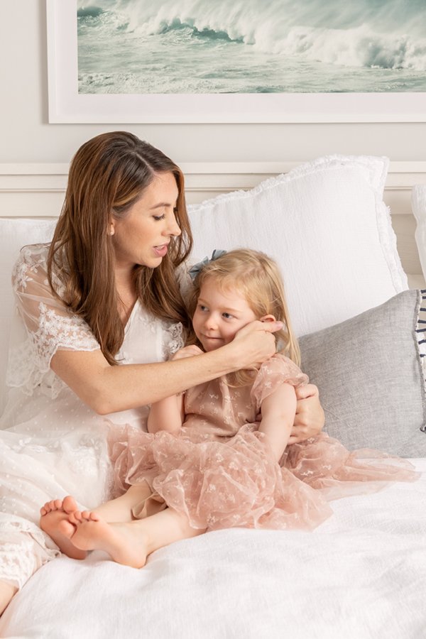  Brunette mom wearing a white lace dress spends quality time with her toddler daughter on her bed. #inhomeportraits #motherdaughter #nicolehawkinsphotography #chatham #newjersey #njfamilyphotographer #firstbornlove #inhomenewbornportraitsession 