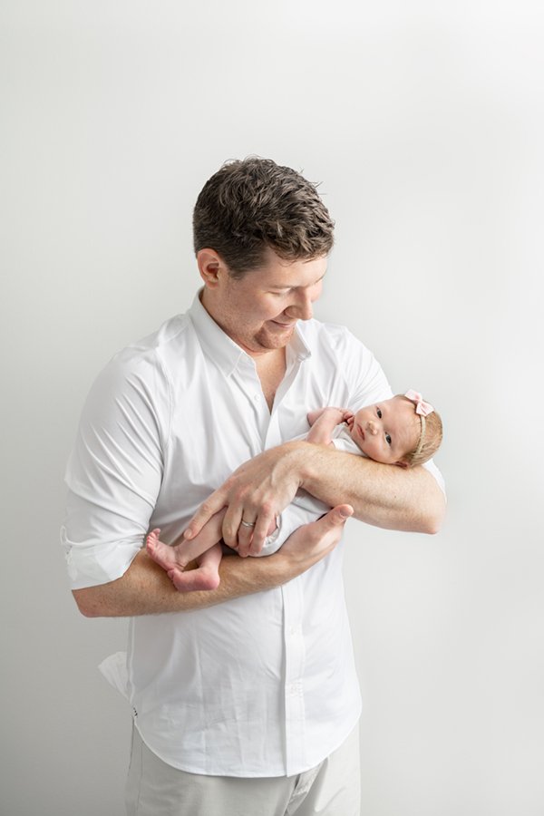  Proud dad wearing a white button down shirt holding his beautiful newborn baby girl in his arms in his New Jersey home. #girldad #njfamilyphotographer #inhomeportraits #nicolehawkinsphotography #chatham #newjersey #nicolehawkinsnewborns #daddysgirl 