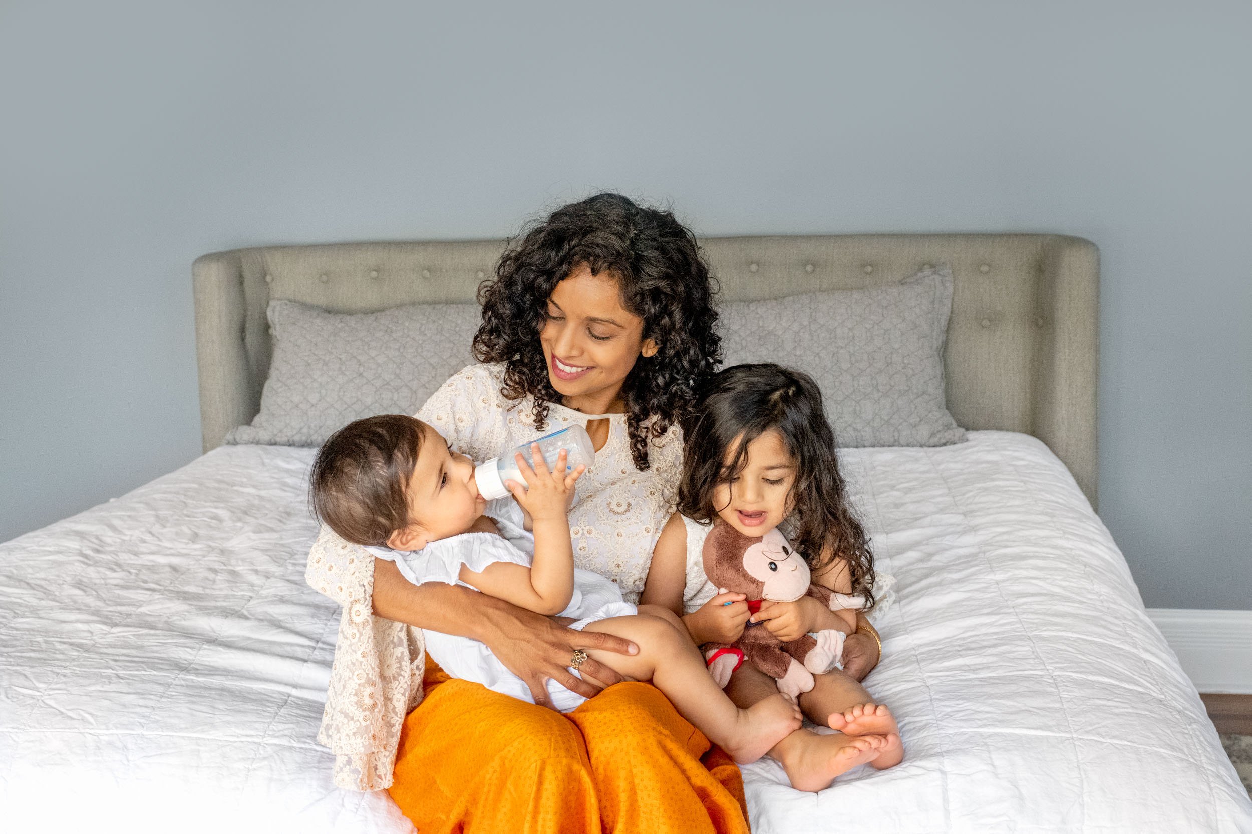  Mom cuddling with her beautiful daughters while one drinks a bottle on her bed in their New Jersey home. #nicolehawkinsfamilies #nicolehawkinsphotography #njfamilyphotographer #newjerseyphotographers #familyportraits #inhomephotographysession 