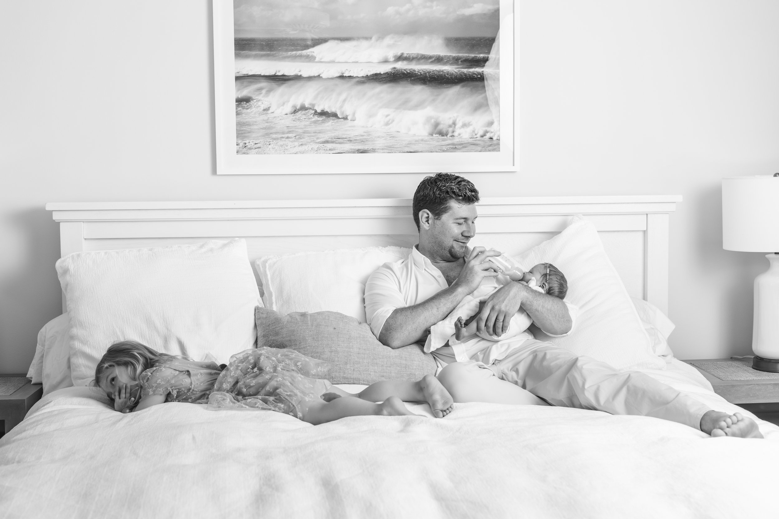  Dad cuddled on his bed feeding his newborn daughter a bottle while his toddler daughter cuddle. #nicolehawkinsnewborns #njfamilyphotographer #chatham #newjersey #newbornfamilyportraits #nicolehawkinsphotography #inhomeportraits #secondtimeparents 