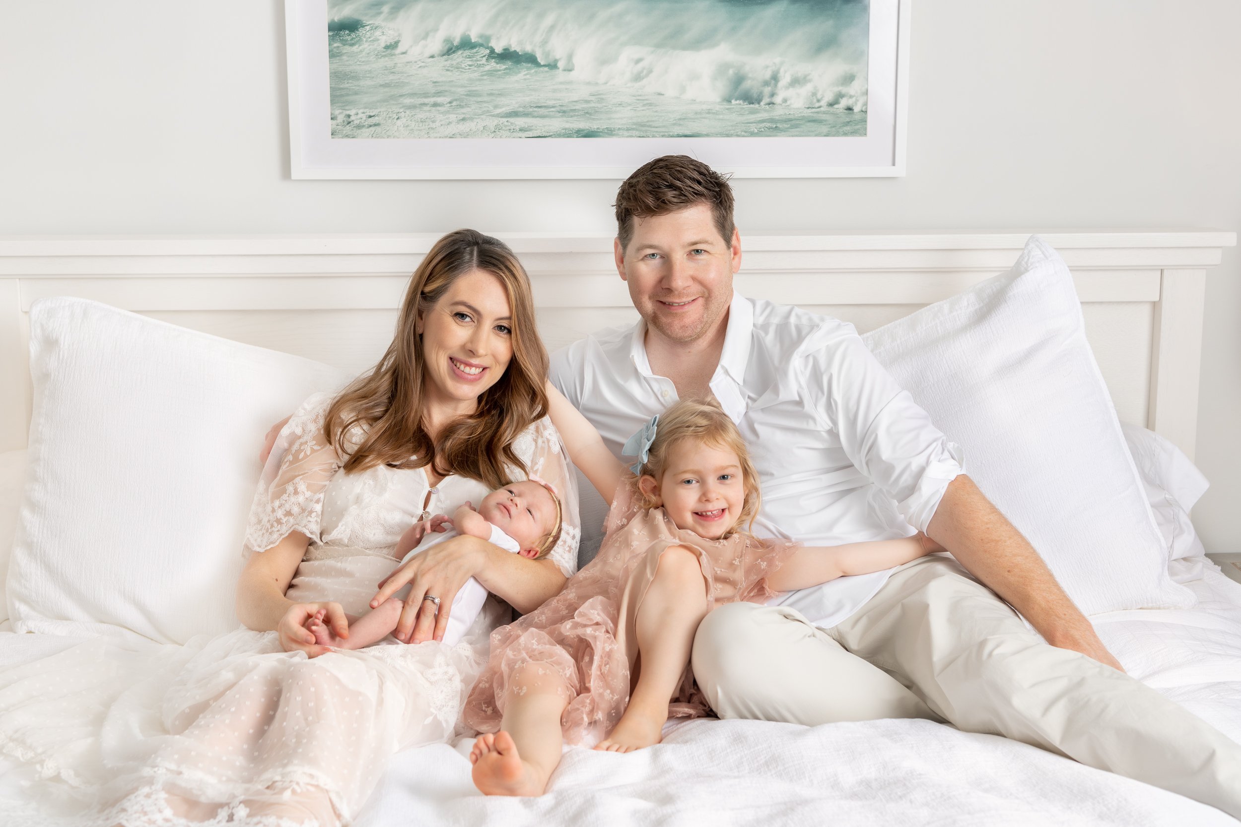  Mother and father sitting on a white bed holding their toddler daughter and newborn baby girl in New Jersey. #newbornsession #njfamiyphotographer #inhomephotography #nicolehawkinsnewborns #nicolehawkinsphotography #secondchild #chatham #newjersey 