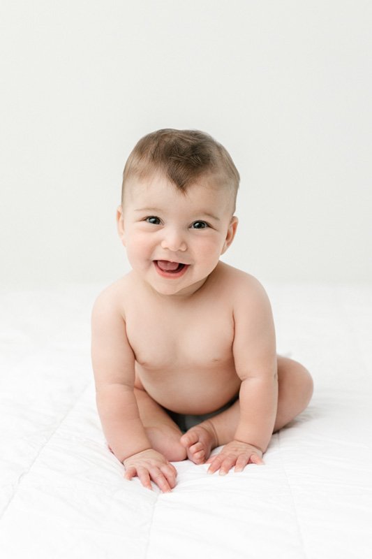  New Jersey Photographer Nicole Hawkins Photography captures a smiling girl sitting on a bed in a studio. New Jersey baby photographers #sixmonthbabyportraits #NJPhotographer #NicoleHawkinsPhotography #NicoleHawkinsBabies #BabyStudioPhotography    