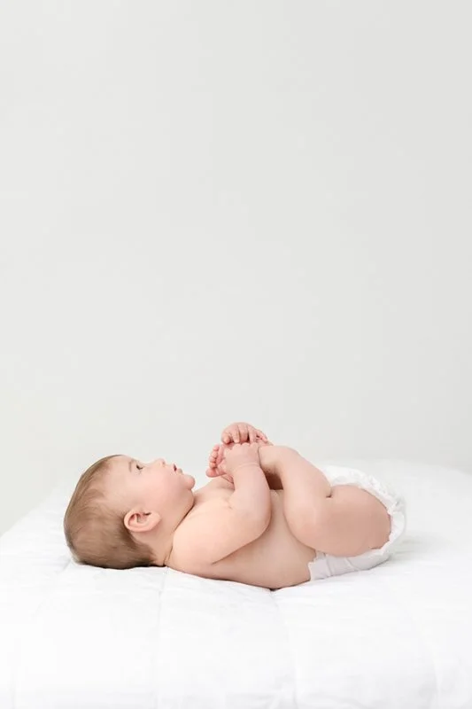  Baby playing with his feet while laying on a bed by Nicole Hawkins Photography. playing baby feet baby girl portraits #sixmonthbabyportraits #NJPhotographer #NicoleHawkinsPhotography #NicoleHawkinsBabies #BabyStudioPhotography    