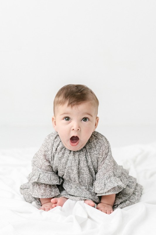  Baby girl with a shocked face wearing a gray ruffle dress by Nicole Hawkins Photography. shocked baby girl face sitter baby studio session #sixmonthbabyportraits #NJPhotographer #NicoleHawkinsPhotography #NicoleHawkinsBabies #BabyStudioPhotography  