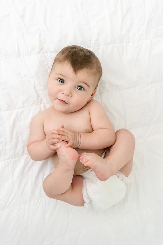  Baby laying down on a bed holding their toes smiling up at their momma by Nicole Hawkins Photography. baby portraits holding his feet #sixmonthbabyportraits #NJPhotographer #NicoleHawkinsPhotography #NicoleHawkinsBabies #BabyStudioPhotography    