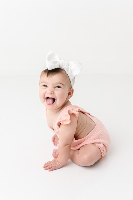  A little girl in a pink strappy onesie and a white bow smiles during a portrait session with Nicole Hawkins Photography. pink baby outfit #sixmonthbabyportraits #NJPhotographer #NicoleHawkinsPhotography #NicoleHawkinsBabies #BabyStudioPhotography   
