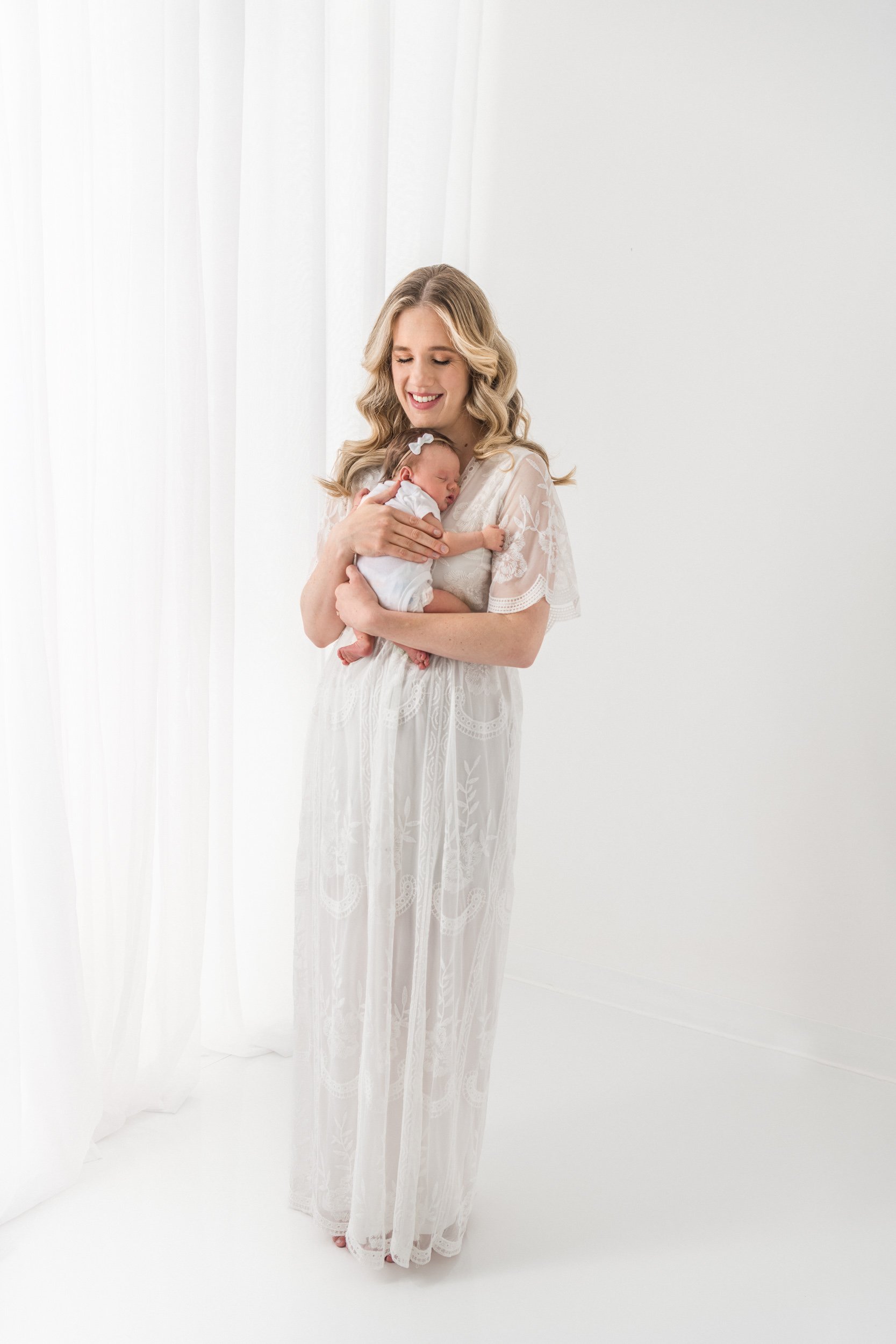  Mother in a white lace dress rocks her baby girl in her arms by Nicole Hawkins Photography. mother and baby motherhood #NicoleHawkinsPhotography #NicoleHawkinsNewborns #StudioPhotography #NewYorkPhotographer #NJphotographers #newborn 