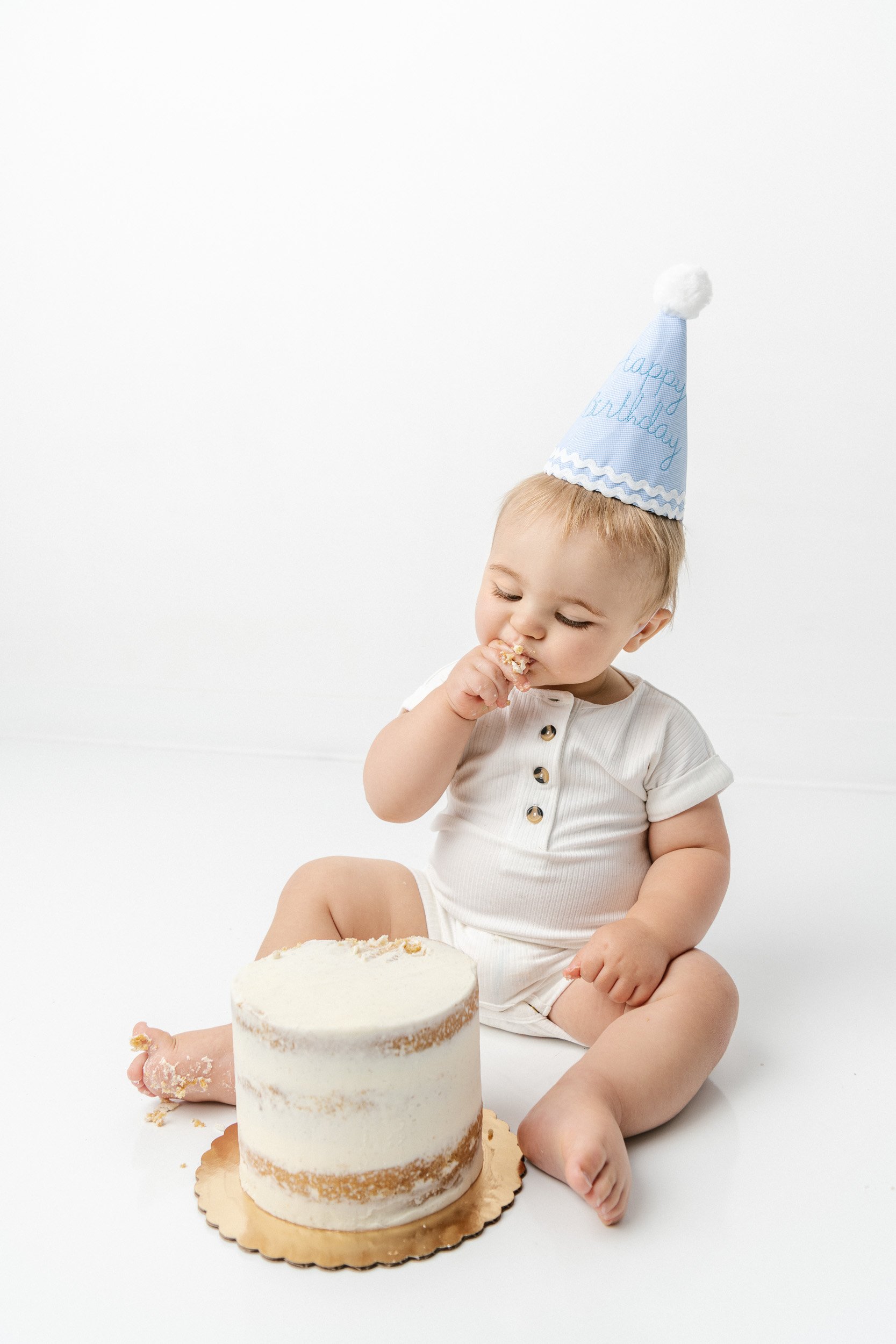  A little boy with a blue party hat in a white romper eating cake by Nicole Hawkins Photography. blue party hat #NicoleHawkinsPhotography #NicoleHawkinsBabies #BirthdayStudioPhotography #smashcake #firstbirthday #studiobabiesandchildren 