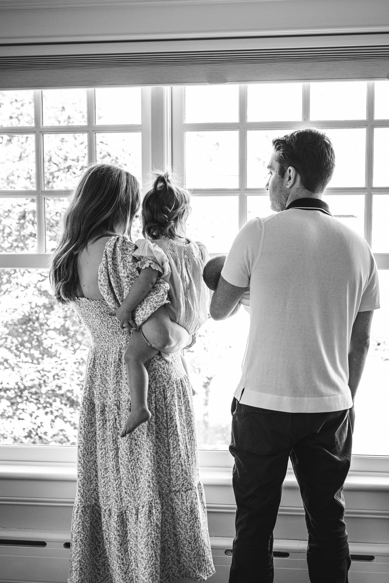  A family of four stands at a window and looks out captured by Nicole Hawkins Photography, a family photographer. fam lifestyle pic #NicoleHawkinsPhotography #InHomeNewborn #JerseyShoreNewborn #MontclairPhotographers #NewbornPhotography #baby #newbor