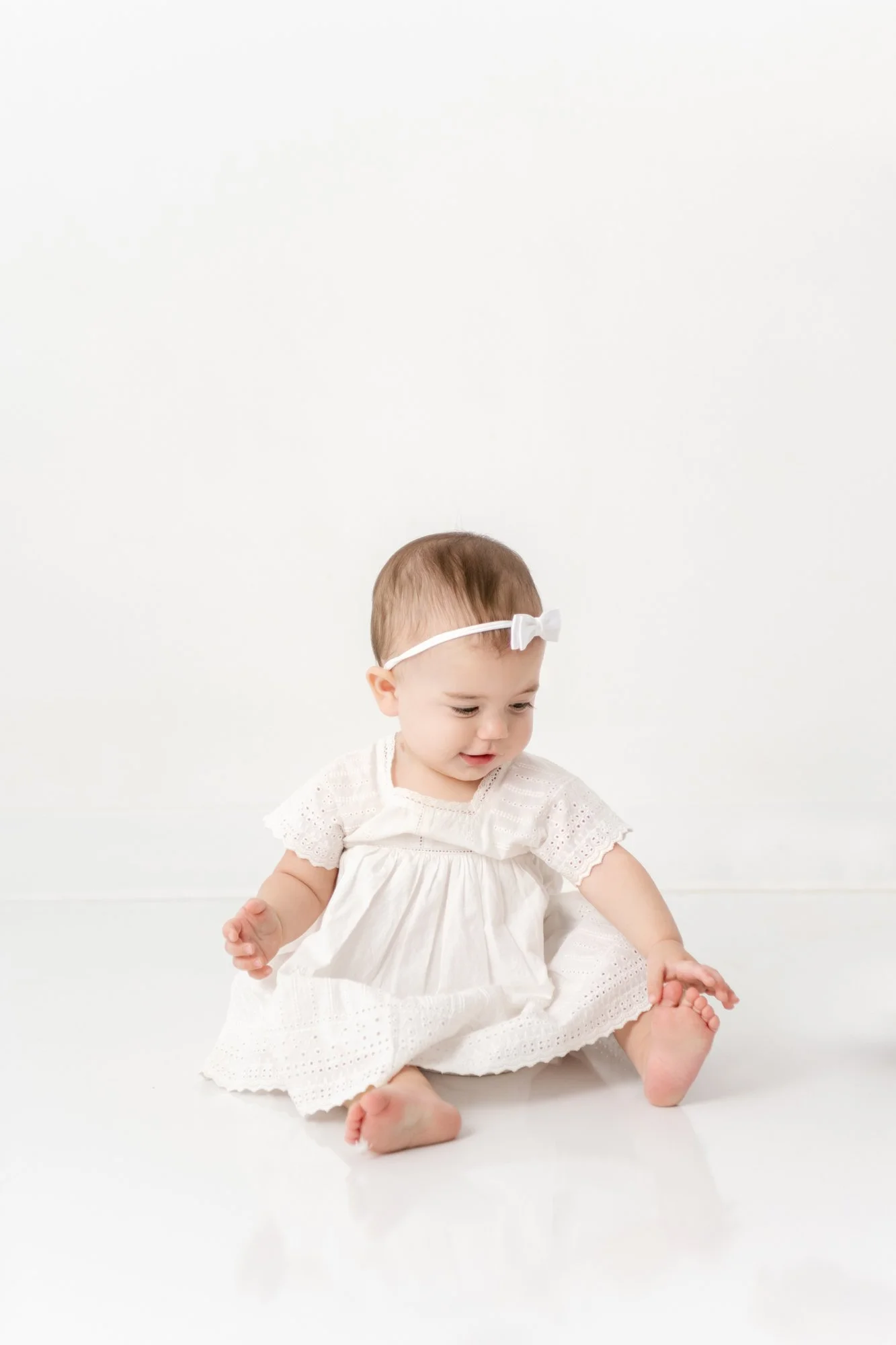 A one-year-old girl in a white dress and bow touching her toes captured by Nicole Hawkins photography. baby girl bow #NicoleHawkinsPhotography #firstbirthday #babyportraits #NJphotographers #lifestylephotography #studiobabies #childrenphotography 