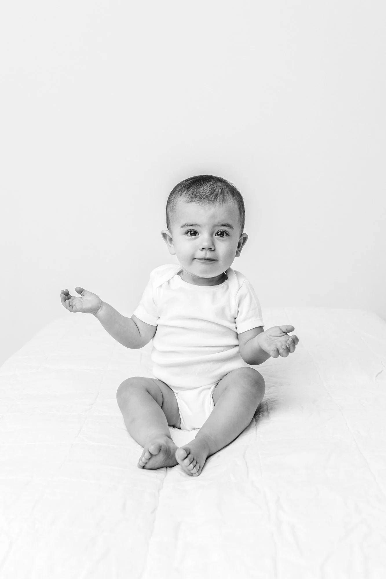 Baby girl holds her hands up while sitting on a white bed captured by Nicole Hawkins Photography. baby on bed #NicoleHawkinsPhotography #firstbirthday #babyportraits #NJphotographers #lifestylephotography #studiobabies #childrenphotography 