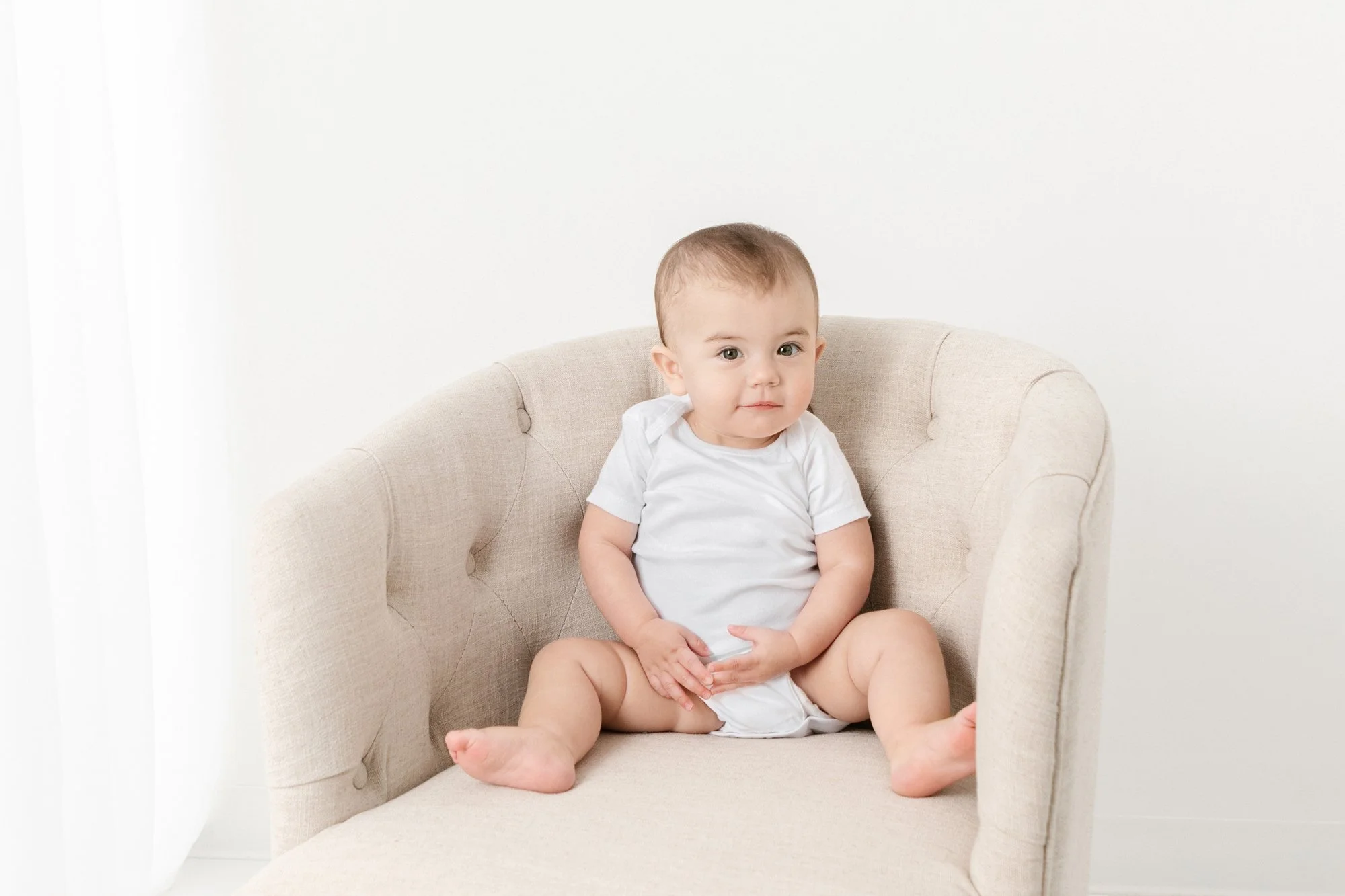  A baby girl siting in a chair in a white onesie captured by Nicole Hawkins Photography. white onesie portraits #NicoleHawkinsPhotography #firstbirthday #babyportraits #NJphotographers #lifestylephotography #studiobabies #childrenphotography 