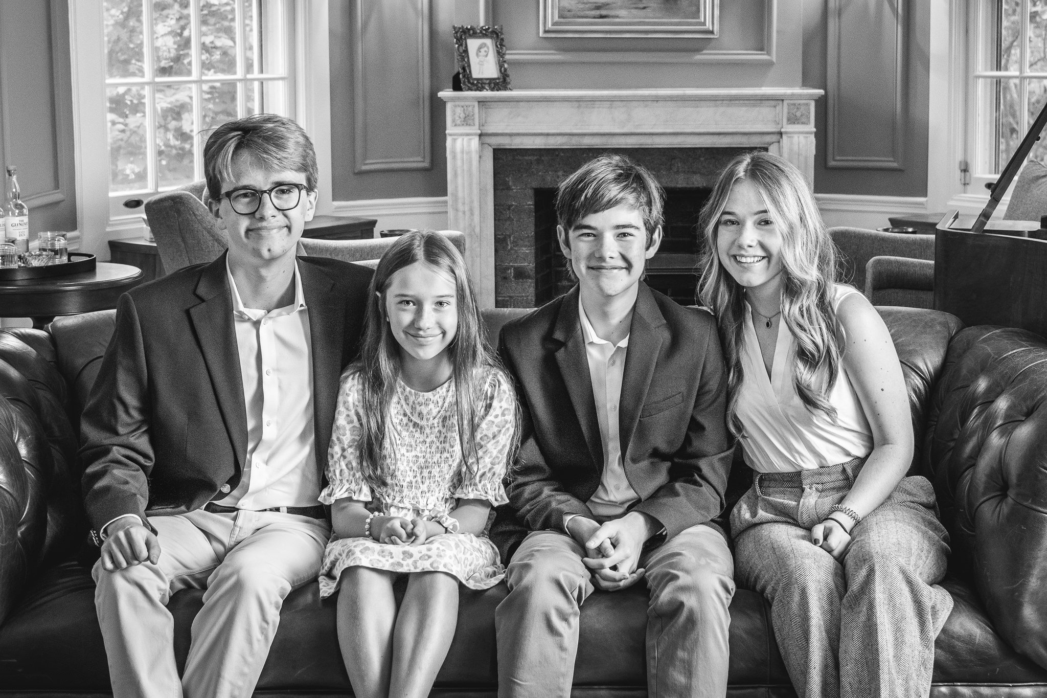  A black and white portrait of four siblings on a leather couch by Nicole Hawkins Photography. fireplace portrait classy portraits #NicoleHawkinsPhotography #NicoleHawkinsFamilyPortrait #NicoleHawkinsSenior #NJPortraits #siblingportrait 