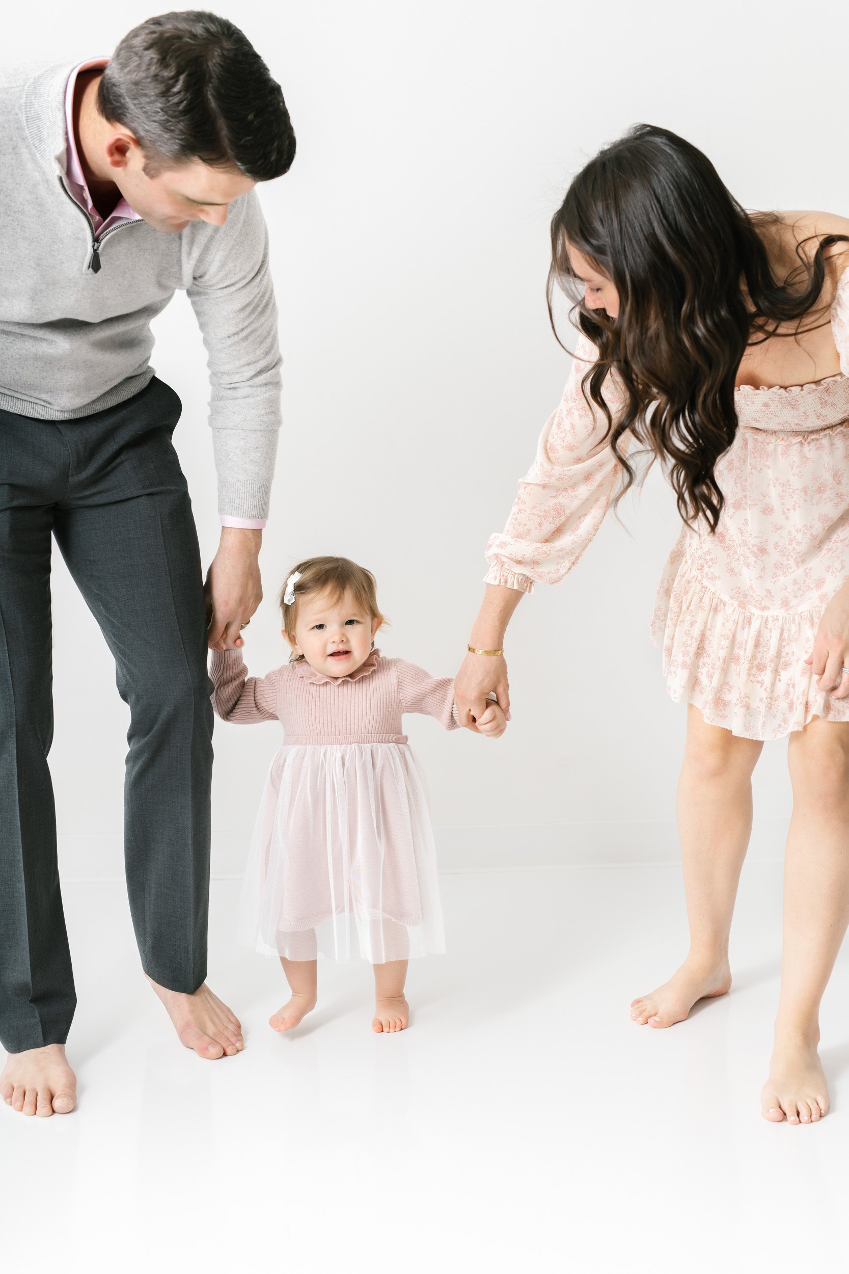  A husband and wife holding their baby girl’s hands helping her walk by Nicole Hawkins Photography. family of three #NicoleHawkinsPhotography #NicoleHawkinsBabies #studiochildren #firstbirthday #studiophotography #girlsbirthdayportraits 