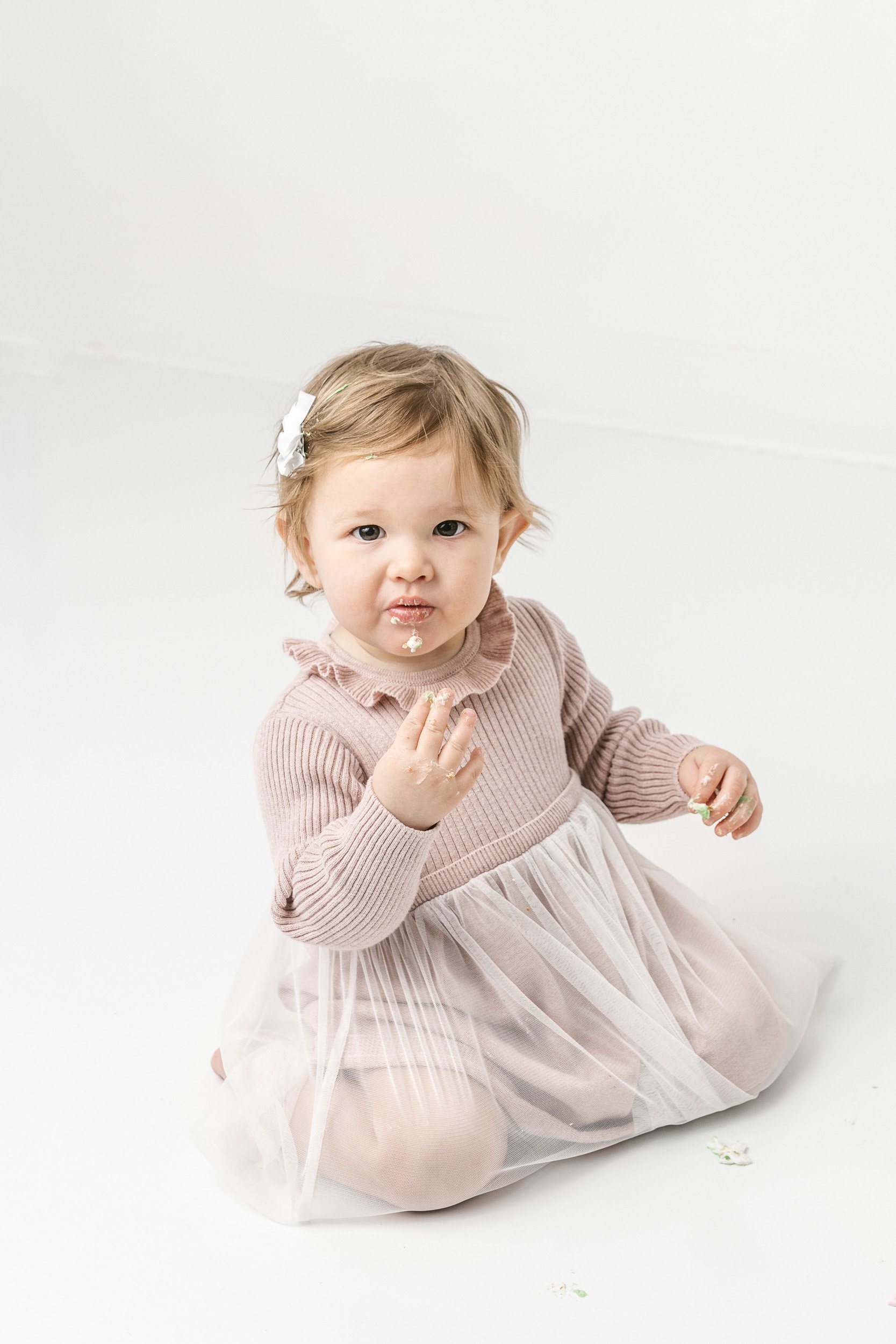  Nicole Hawkins Photography captures a baby girl tasting birthday cake in a white bright studio. studio smash cake #NicoleHawkinsPhotography #NicoleHawkinsBabies #studiochildren #firstbirthday #studiophotography #girlsbirthdayportraits 
