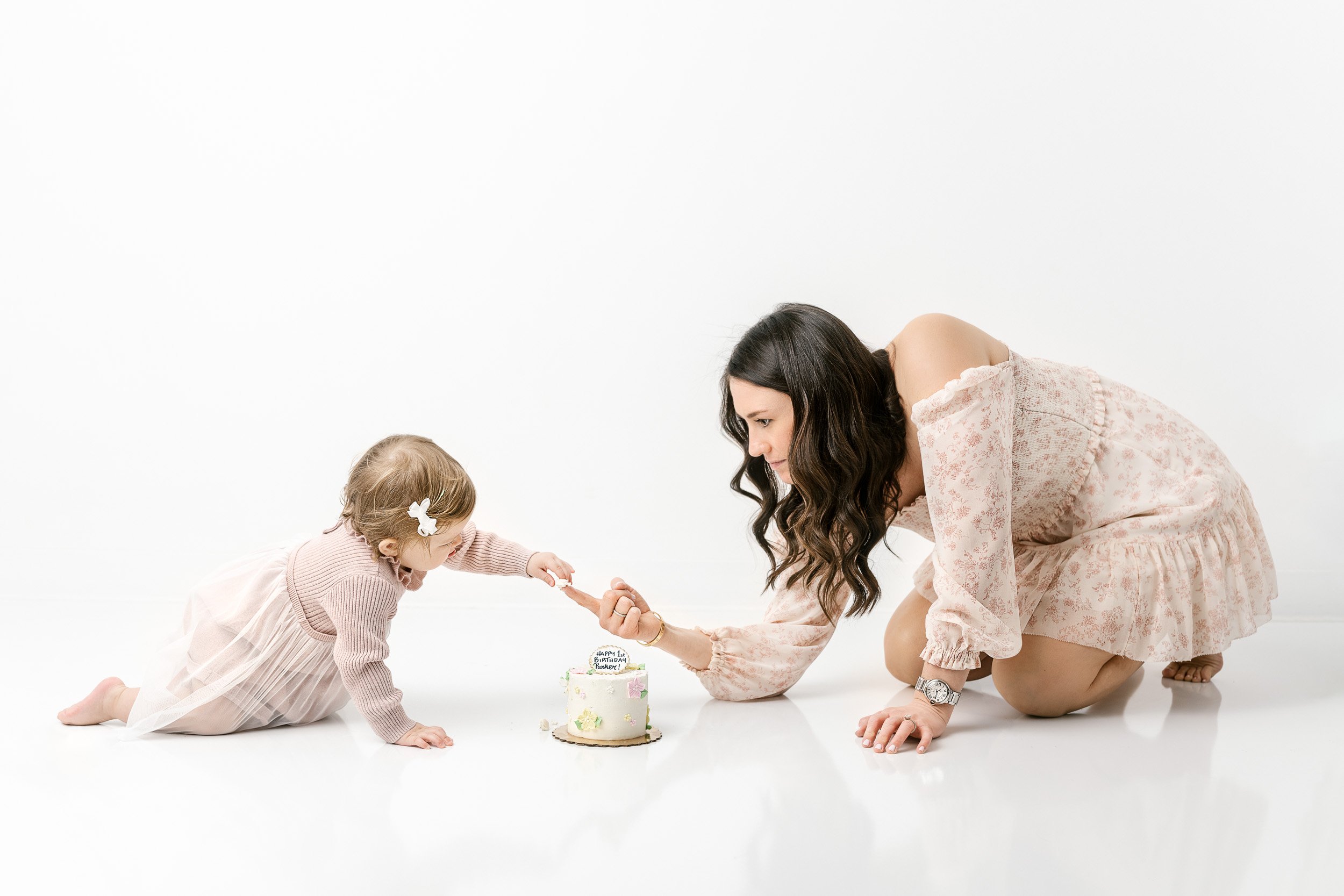  A mother helps her baby girl try her birthday cake during a baby portrait session by Nicole Hawkins Photography. sharing cake #NicoleHawkinsPhotography #NicoleHawkinsBabies #studiochildren #firstbirthday #studiophotography #girlsbirthdayportraits 