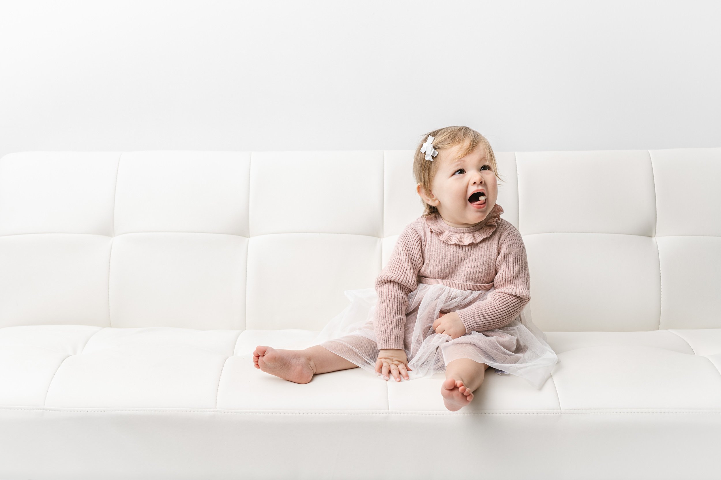   Baby studio photographer Nicole Hawkins Photography captures a baby sitting on a white couch and making silly faces. baby #NicoleHawkinsPhotography #NicoleHawkinsBabies #studiochildren #firstbirthday #studiophotography #girlsbirthdayportraits 